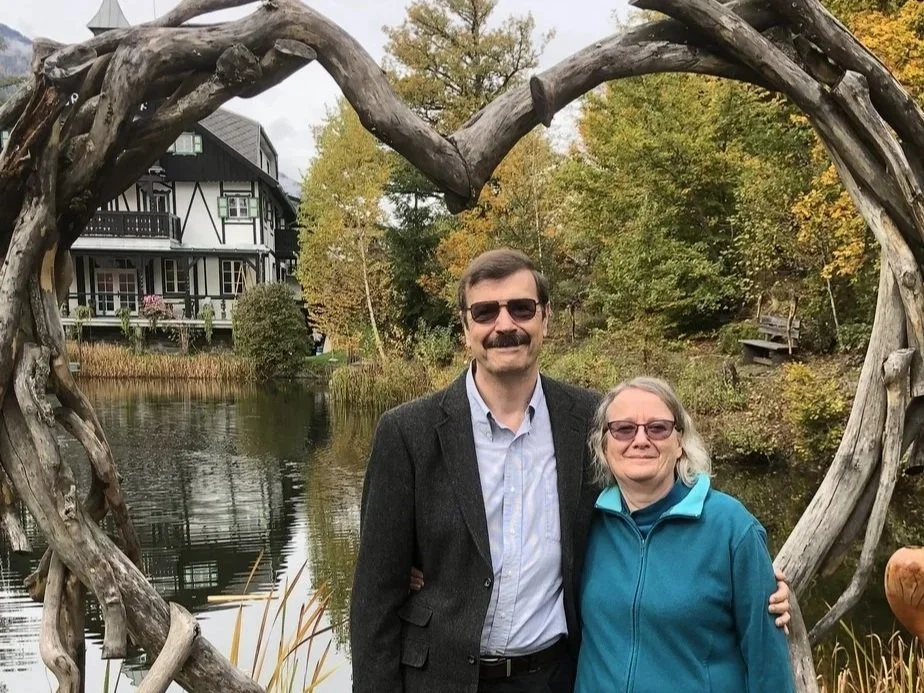 A man and woman standing together under a heart-shaped wooden arch near a pond with a house and trees in the background, during autumn.