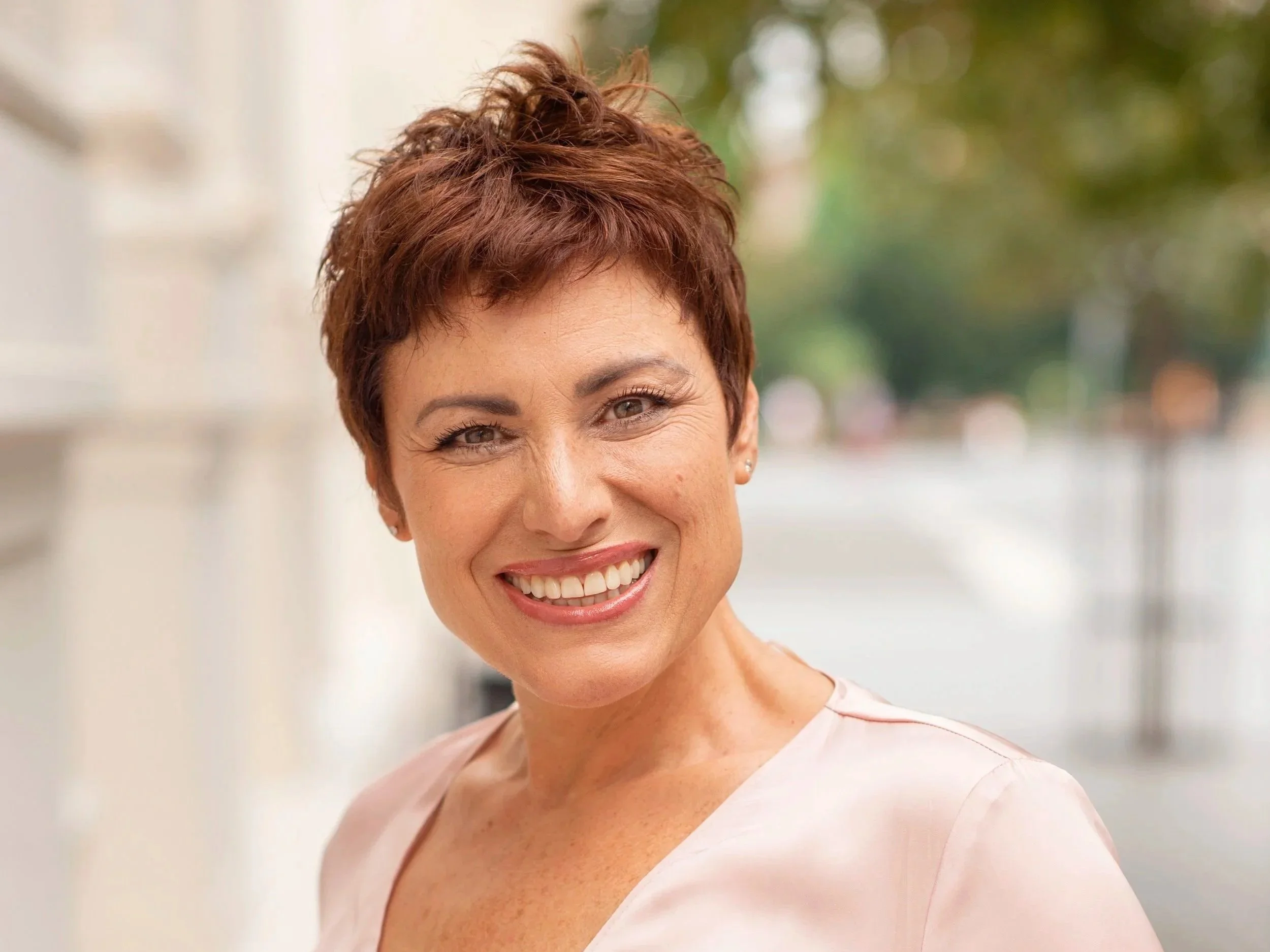 Smiling woman with short, wavy, reddish-brown hair outdoors, background blurred with green and natural colors.