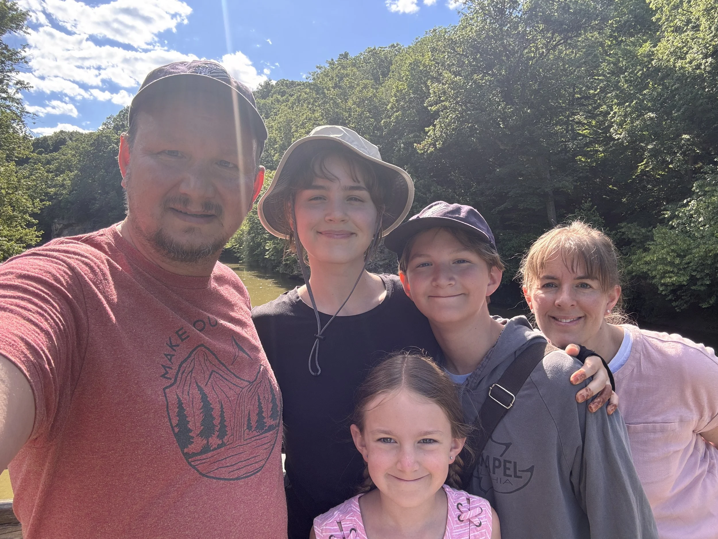 A group photo of four people outdoors on a sunny day, with a blue sky and green trees in the background. The group includes one man and three children, all smiling and dressed casually, with some wearing hats.