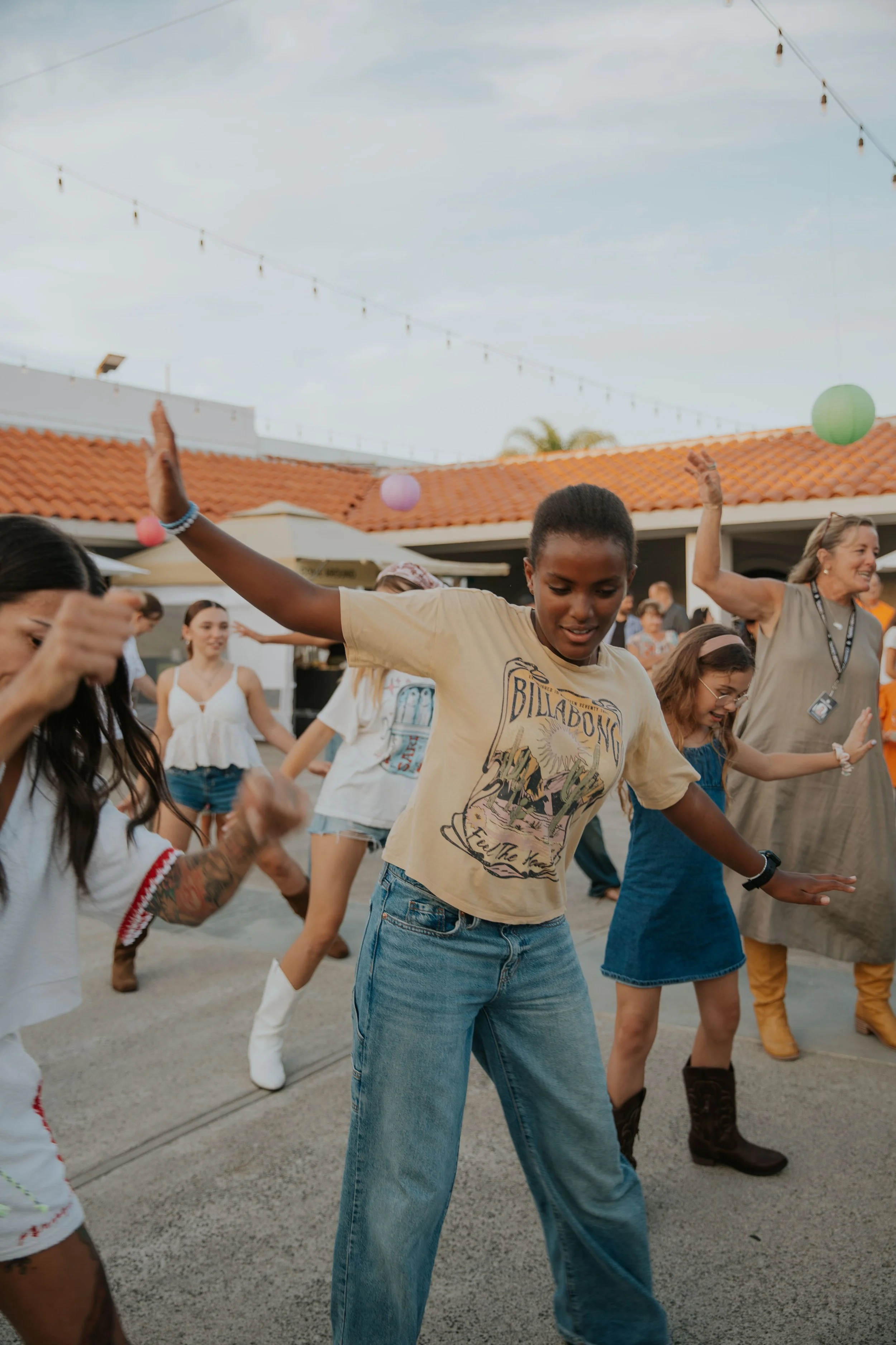 A diverse group of people dancing outdoors under string lights on a cloudy day, some wearing boots and casual summer clothing.