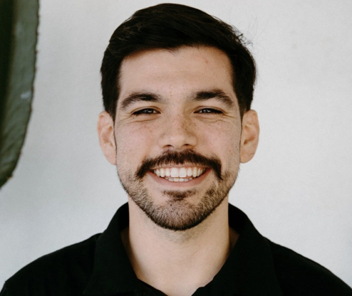 Close-up of a young man with short dark hair, a beard, and a mustache, smiling showing his teeth, wearing a black shirt, standing in front of a light-colored wall.