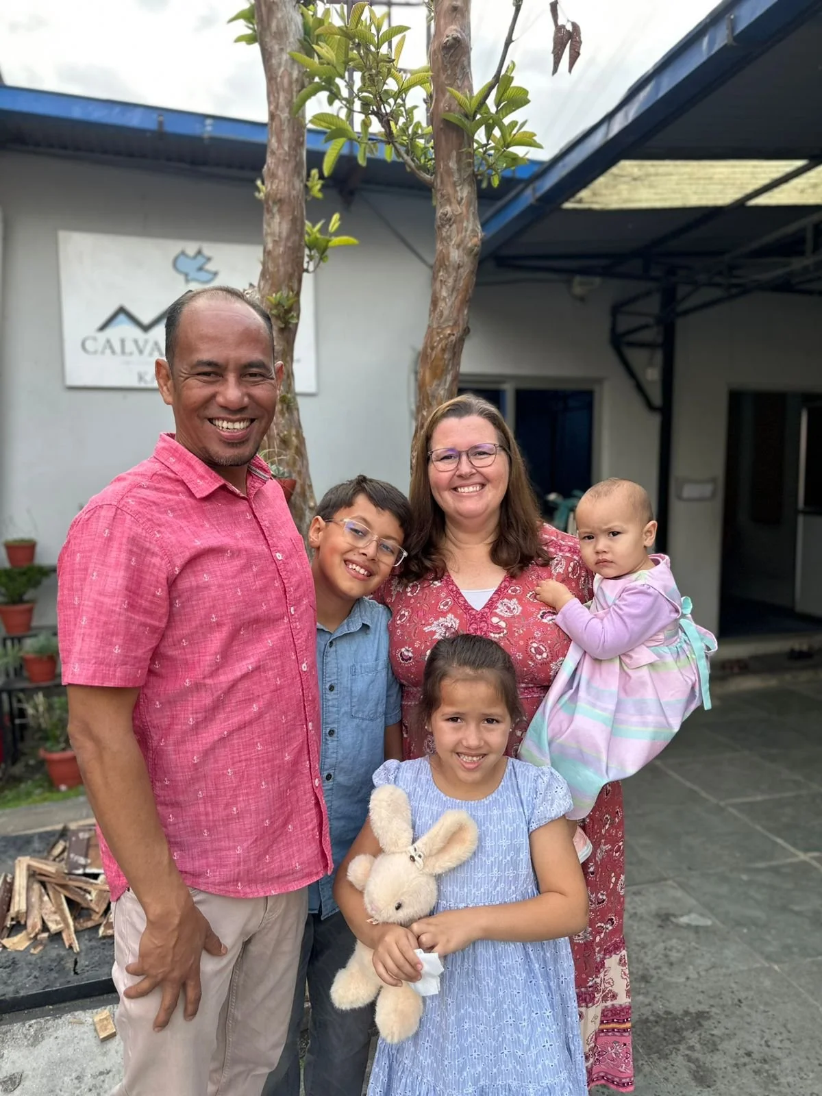 A family of six smiling outdoors in front of a building with a sign, including two adults, three children, and a toddler holding a stuffed bunny.