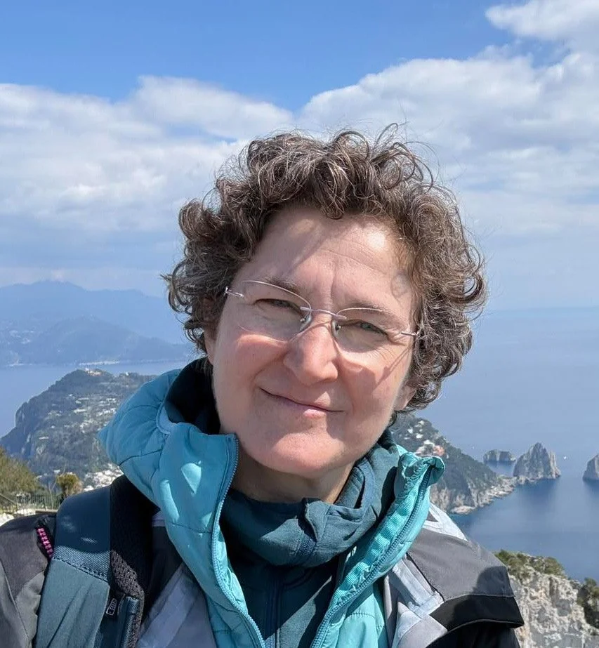 A woman with short curly hair and glasses taking a selfie outdoors with mountains, water, and rocky formations in the background on a bright day.