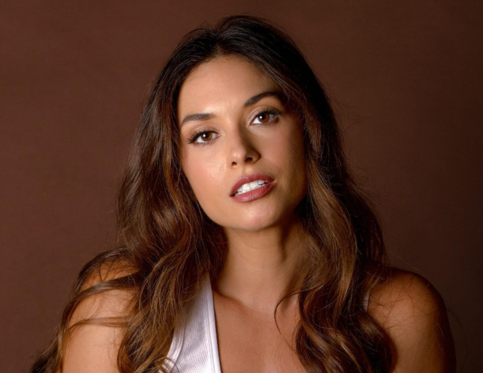 Justene Alpert with long, wavy brown hair looking directly at the camera against a brown background.