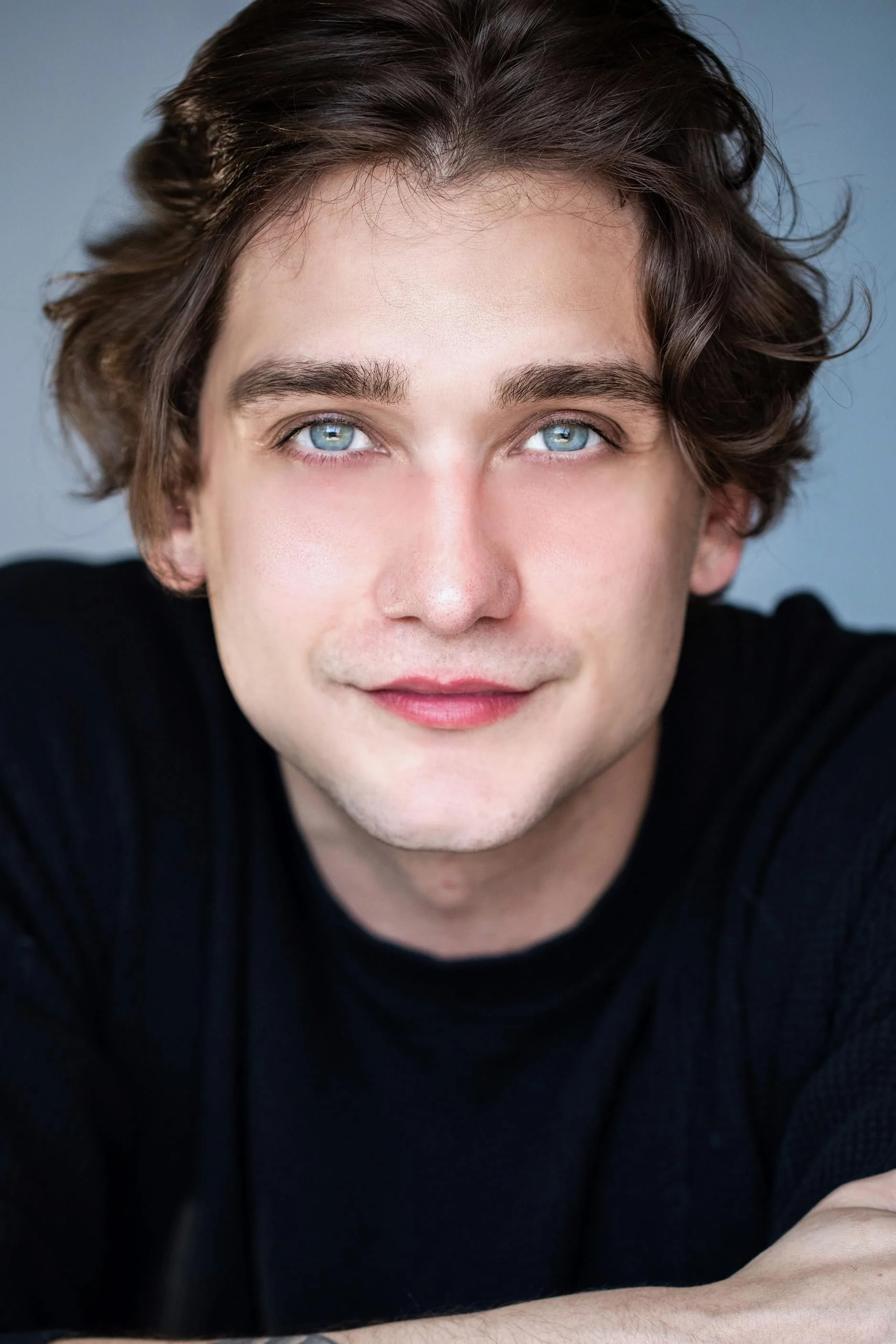 Close-up of a young man with wavy brown hair and blue eyes, wearing a black shirt, leaning forward with a slight smile. Cody Saingnue.