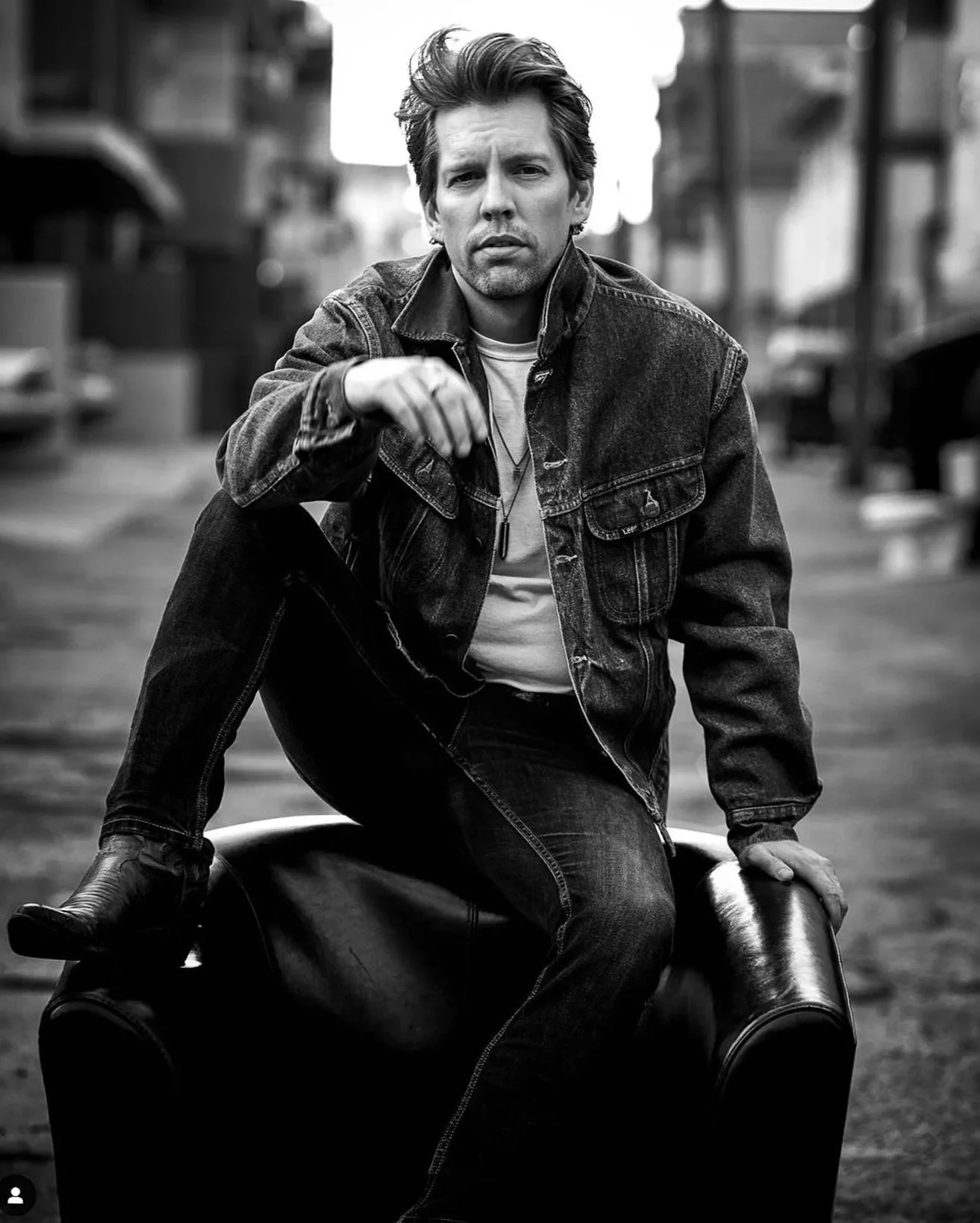 A black and white photo of a man sitting on a motorcycle in an urban street setting. The man is wearing a denim jacket and has a serious expression, with one hand resting on the motorcycle's seat and the other reaching out toward the camera.