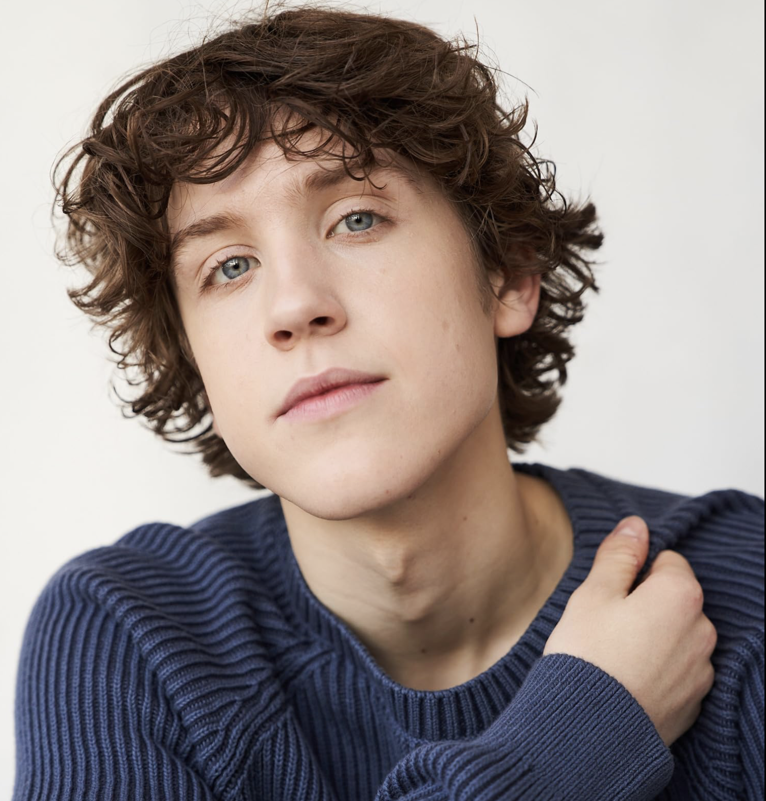 Tanner Hagen with curly short brown hair and blue eyes wearing a navy blue ribbed sweater, touching their shoulder while looking at the camera against a white background.
