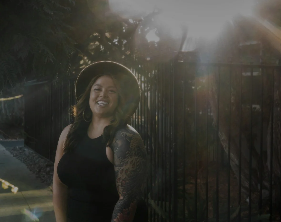 Jamie Hann, Temple Wellness founder and San Diego hair extension & holistic wellness specialist, smiling outdoors in a black sleeveless top and wide-brimmed hat by a metal fence with sunlight filtering through trees
