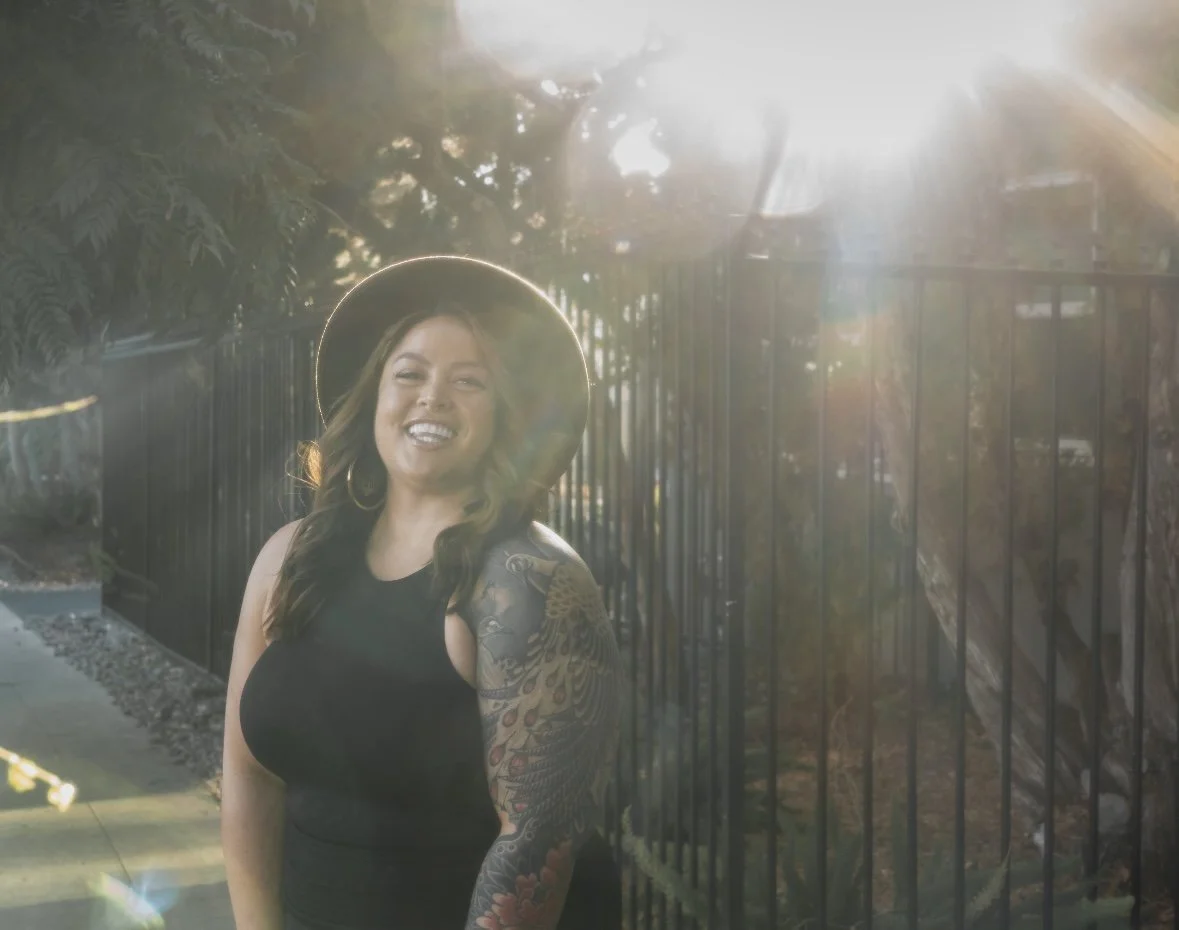 Jamie Hann, Temple Wellness founder and holistic wellness guide, smiling outdoors in San Diego in a black sleeveless dress and wide-brimmed hat by a black fence with sunlight lens flare