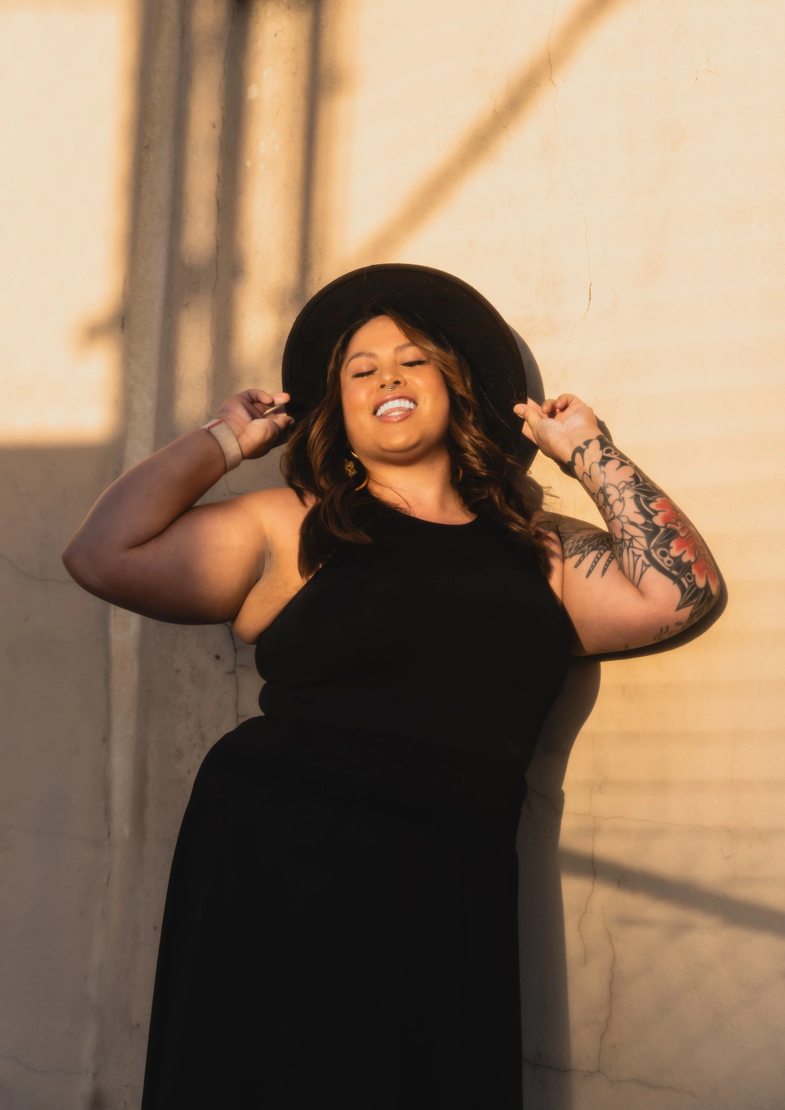 Jamie Hann, Temple Wellness founder and holistic wellness guide, smiling in a black sleeveless dress and wide-brimmed hat, holding her hat and leaning against a beige wall with a decorative fence-shadow pattern