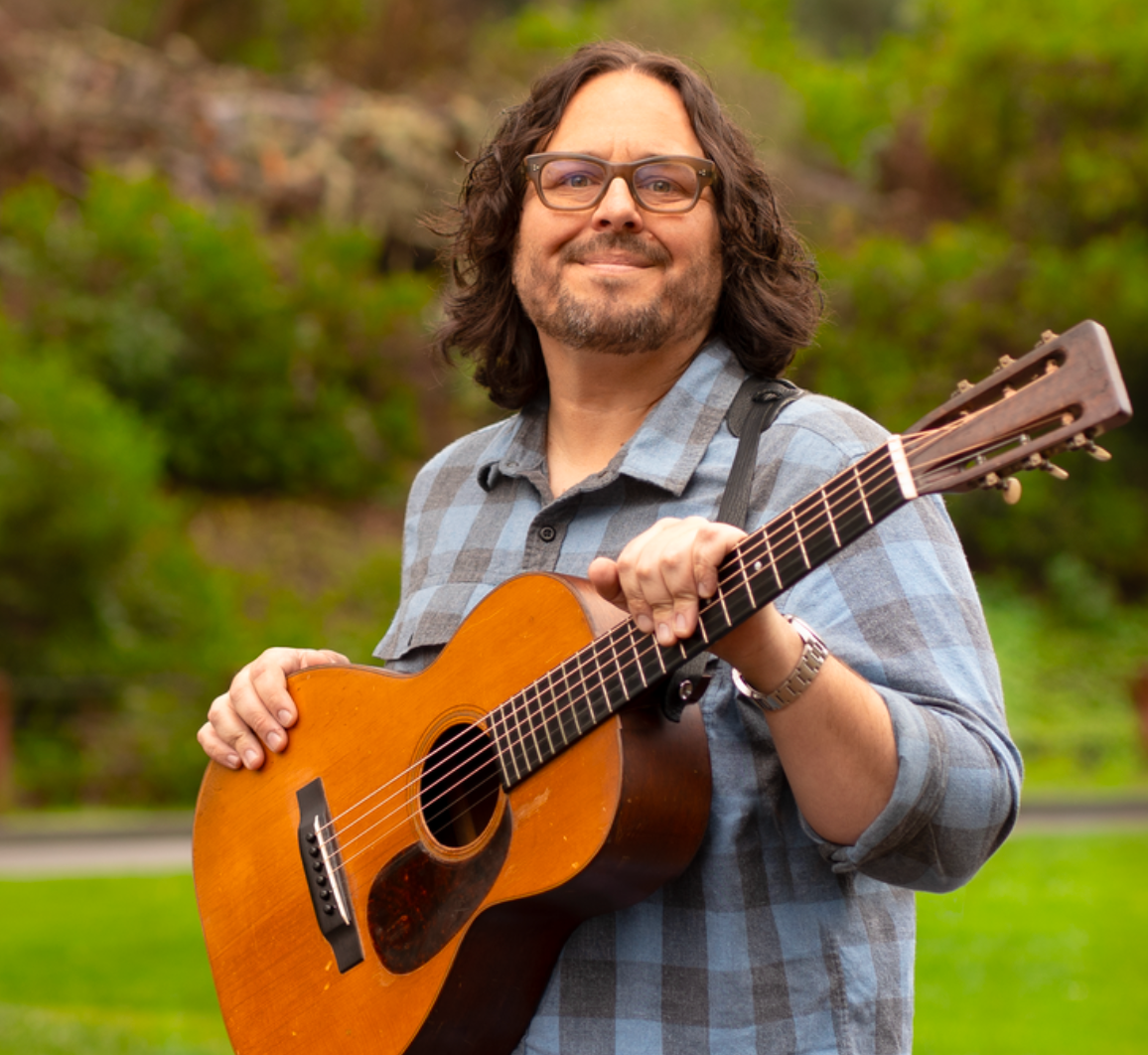 A man with long curly hair, glasses, and a beard playing an acoustic guitar outdoors with greenery in the background.