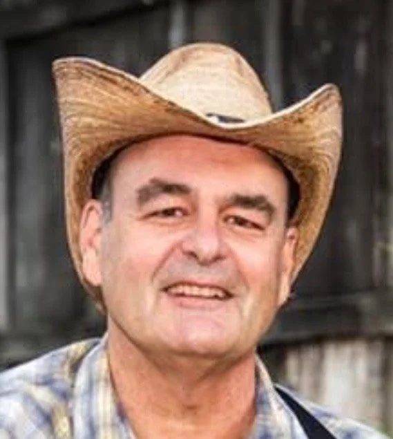 Mike Wilhoyte. A middle-aged man wearing a cowboy hat and plaid shirt, smiling outdoors in front of a wooden fence. Legendary Bluegrass guitarist.