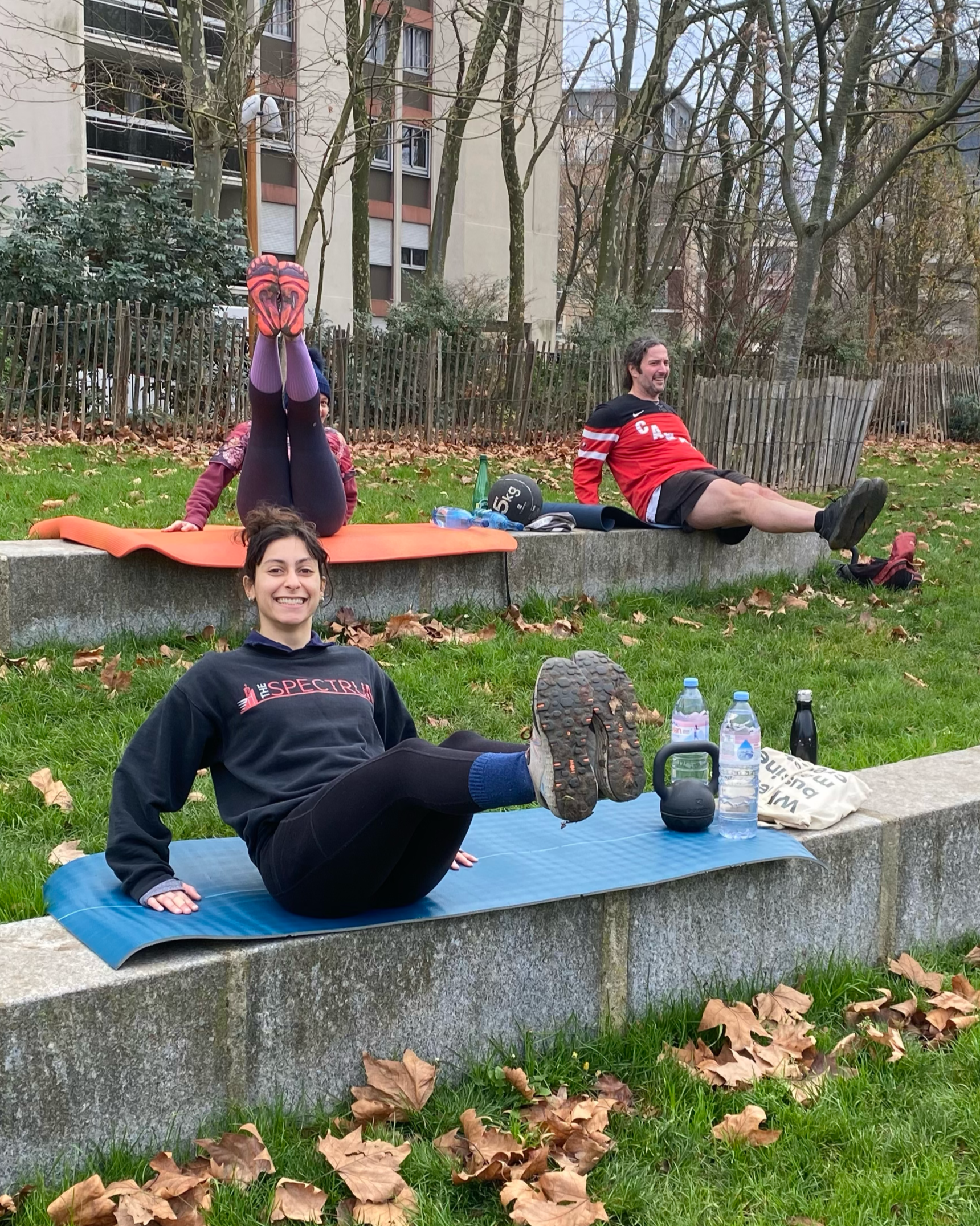 People exercising outdoors on a park bench in autumn, with fallen leaves on the grass, a woman on a mat stretching her legs upwards, a man reclining and smiling, and a woman sitting in front smiling, surrounded by trees and residential buildings.