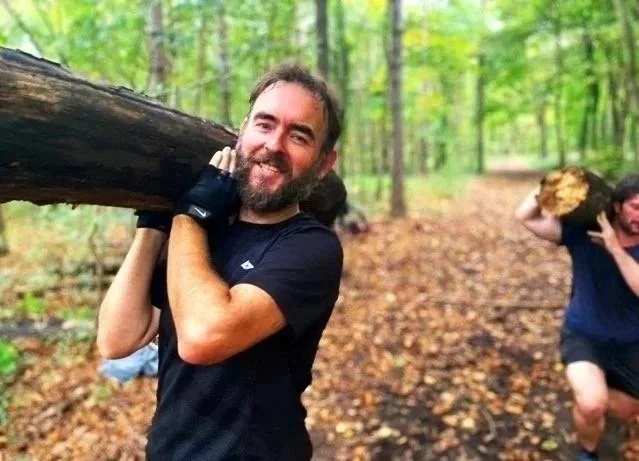 Two men carrying logs in a forest, smiling and walking along a leaf-covered trail.