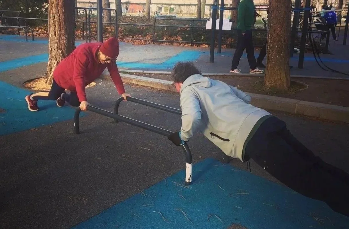 Two people exercising on parallel bars at a park during the daytime, with other park visitors in the background.