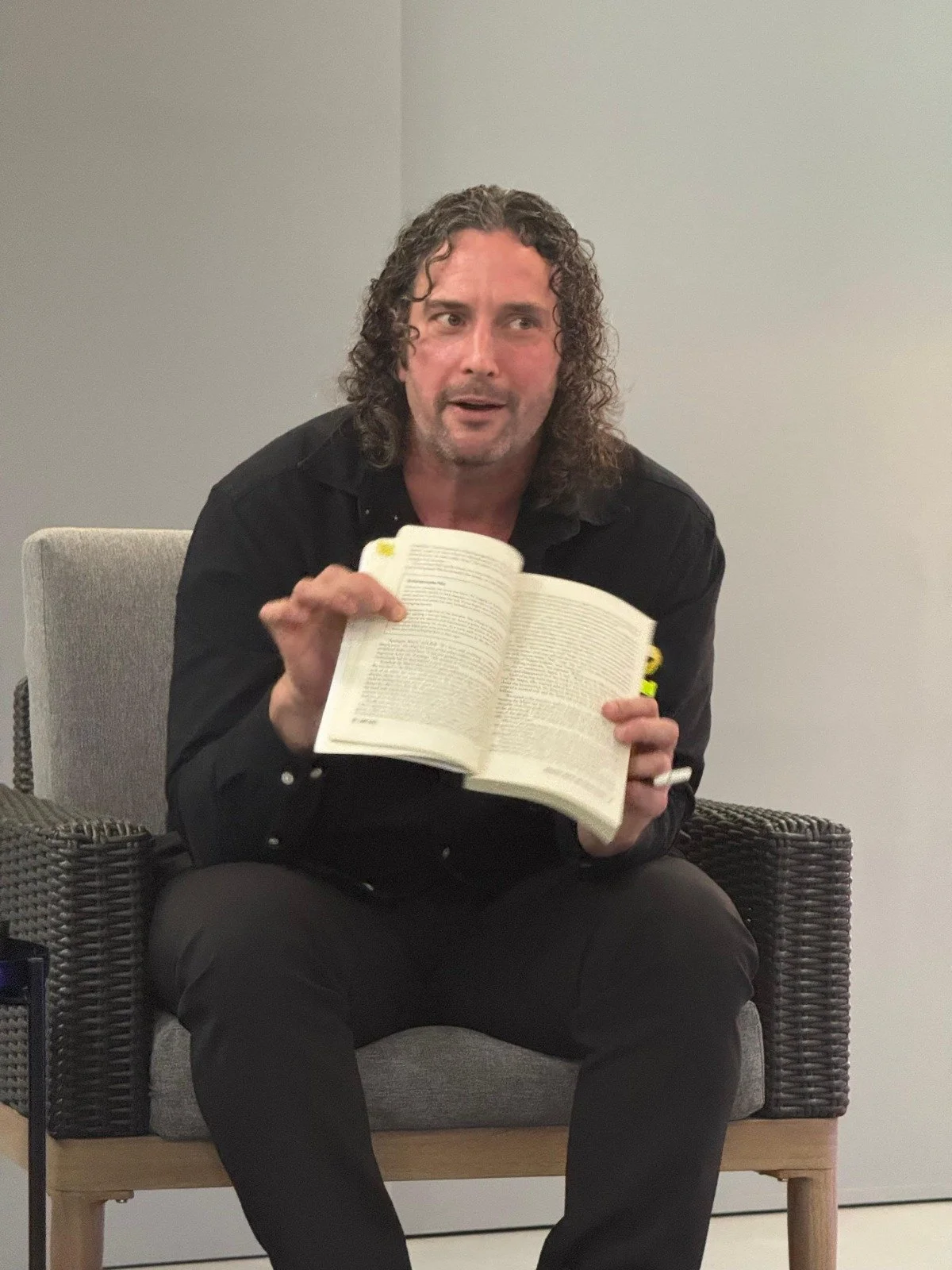 A man with curly dark hair, wearing a black shirt, sitting on a gray chair, holding an open book and looking slightly confused or thoughtful.