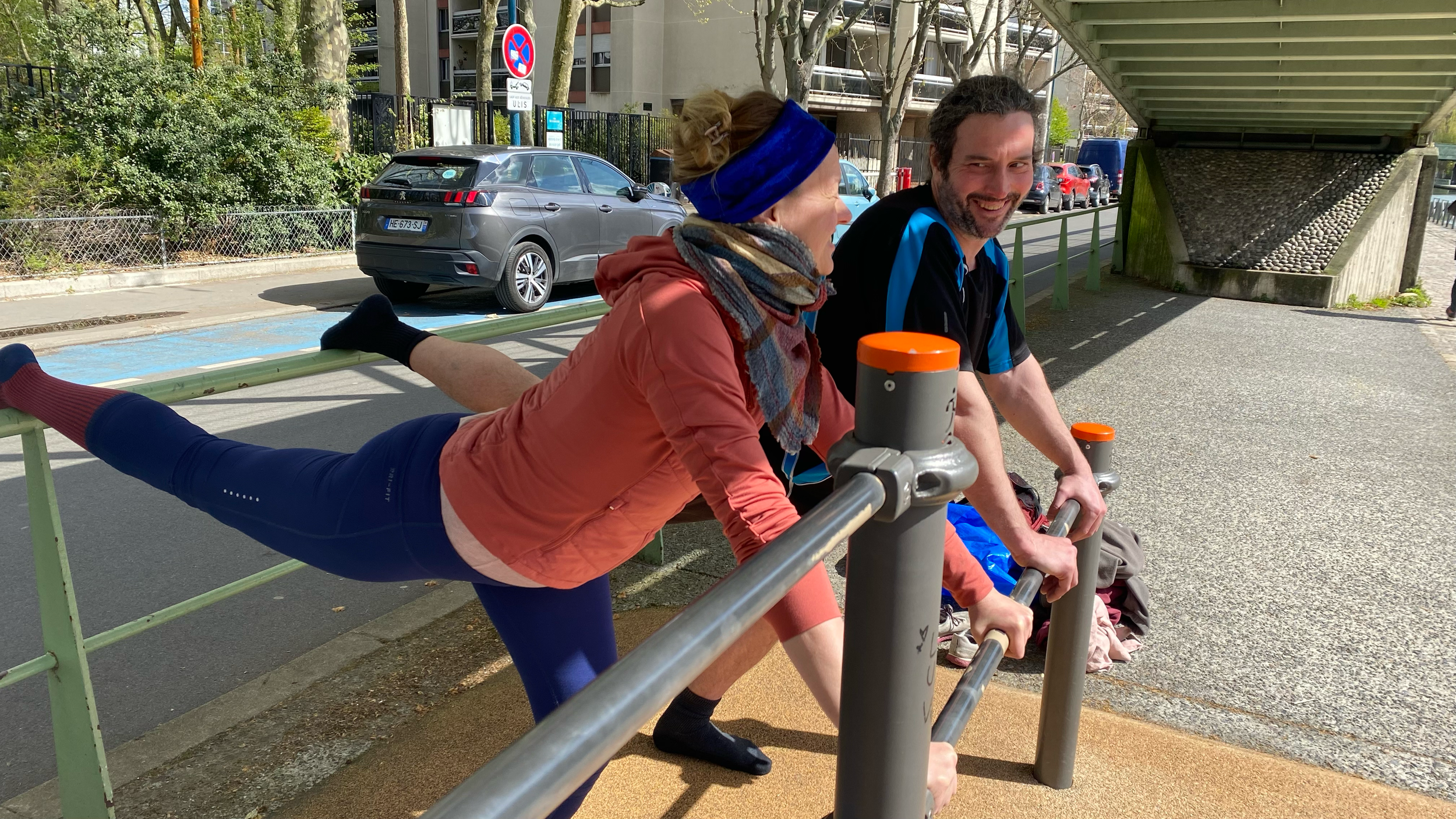 A woman wearing a headband and a scarf doing a split stretch on a railing next to a man sitting and smiling under a bridge.