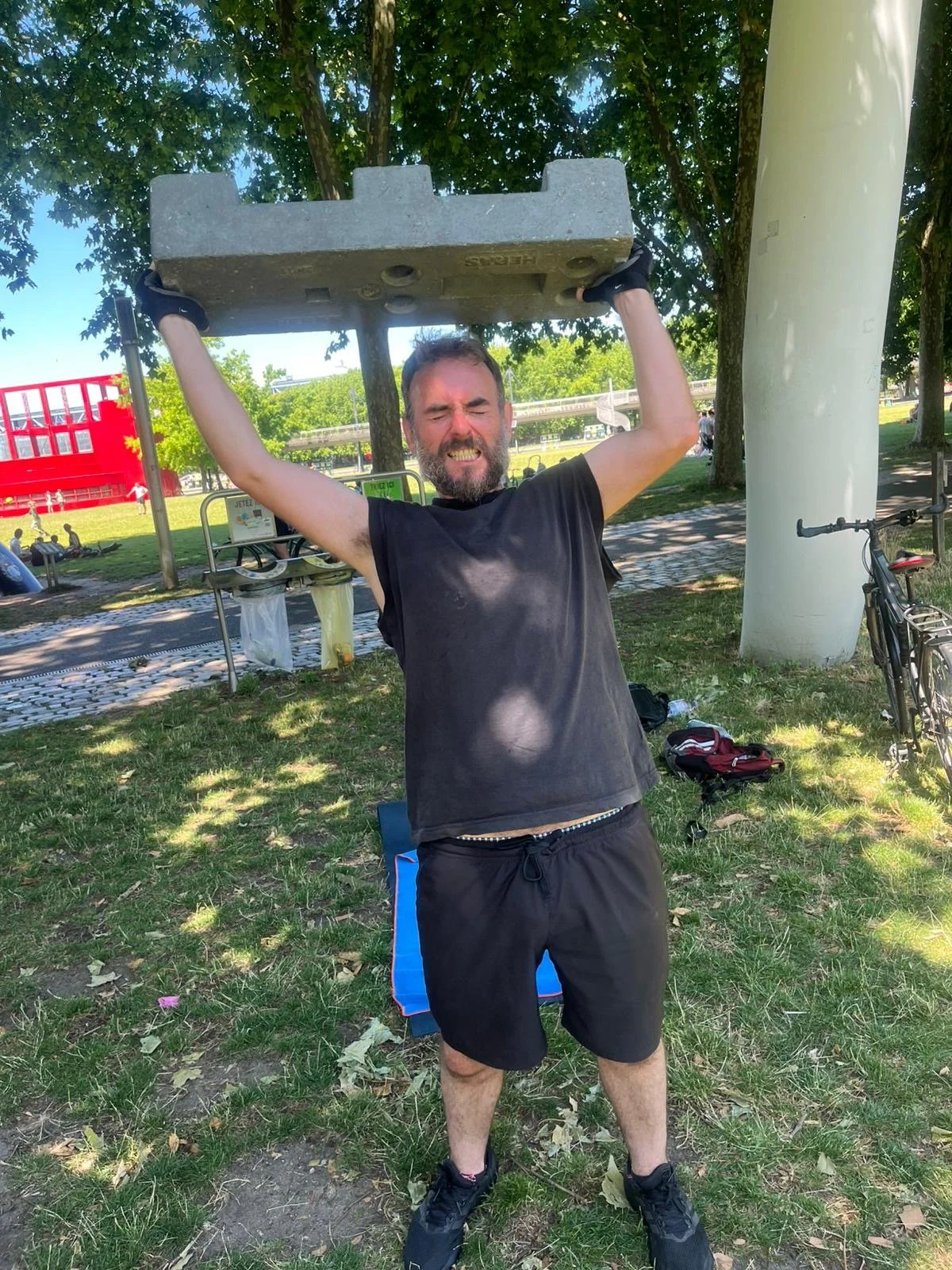A man lifting a concrete block above his head outdoors in a park on a sunny day, with trees and recreational structures in the background.