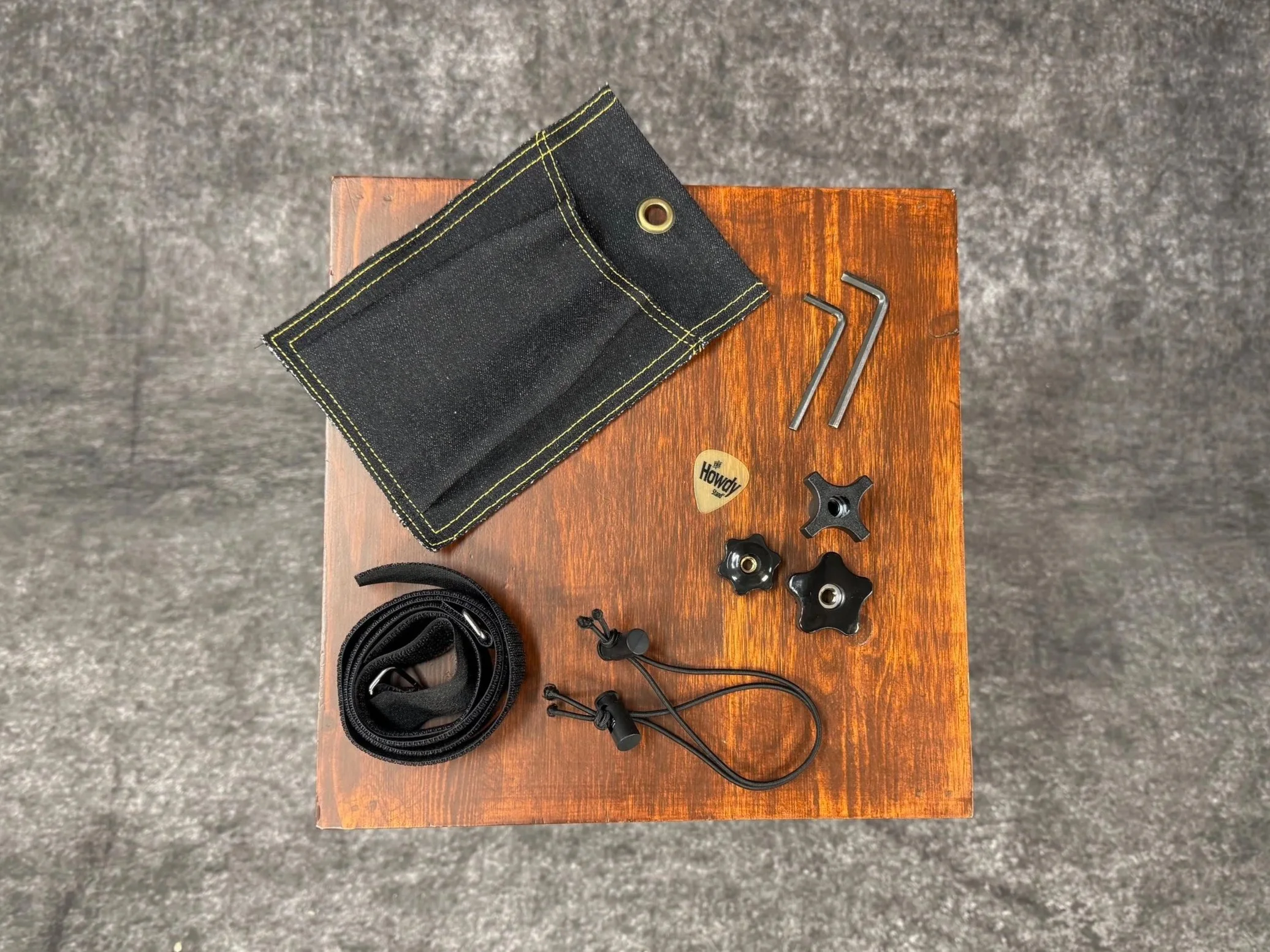A collection of mountain dulcimer stand accessories on a small wooden table, including a black demin pouch, two Allen wrenches, several knobs, two small bungee straps, a dulcimer pick,, and a coiled black hood and loop straps.