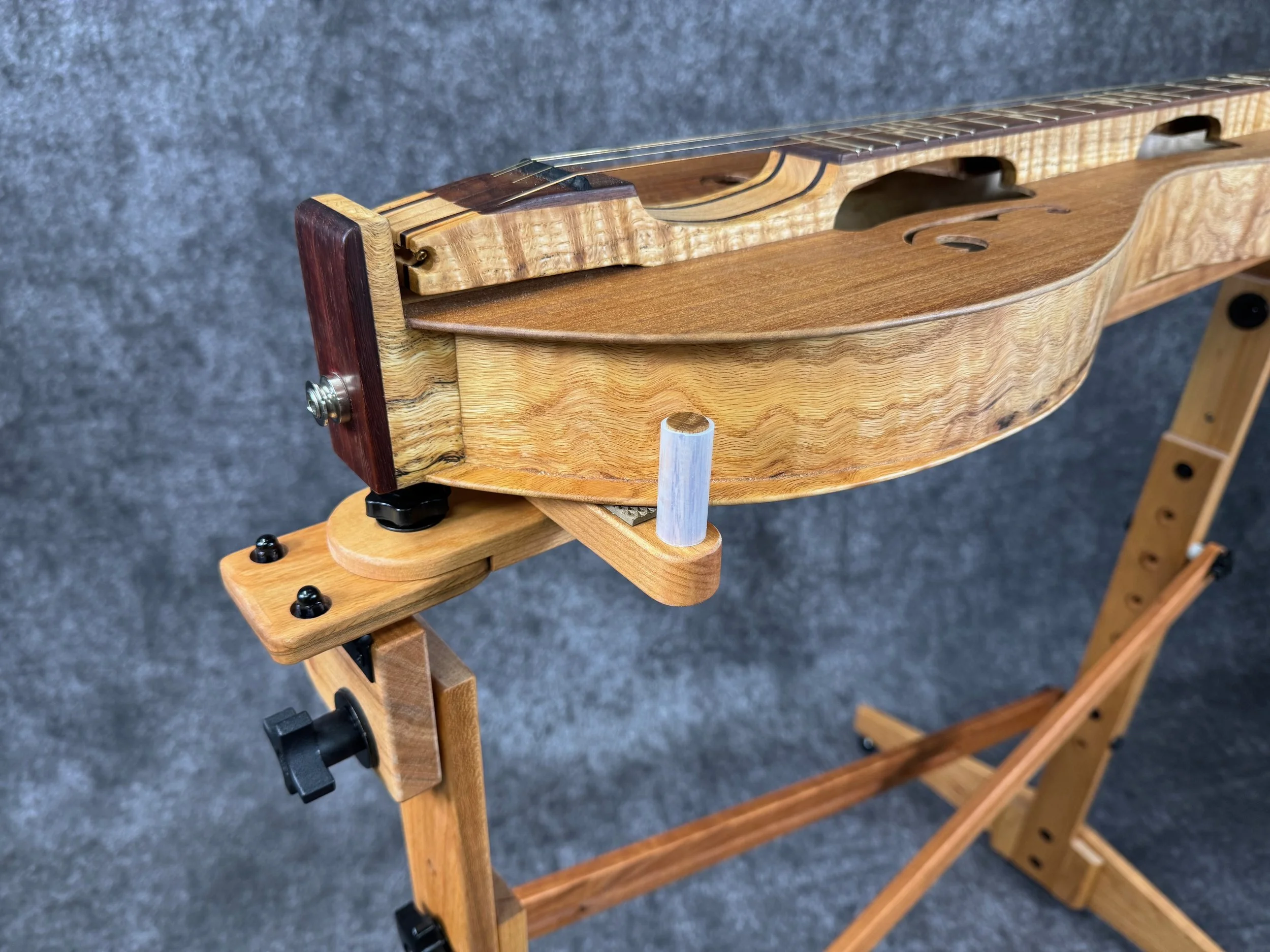 A side rear view of a mountain dulcimer on a wooden stand, showing the tailblock and strap button.