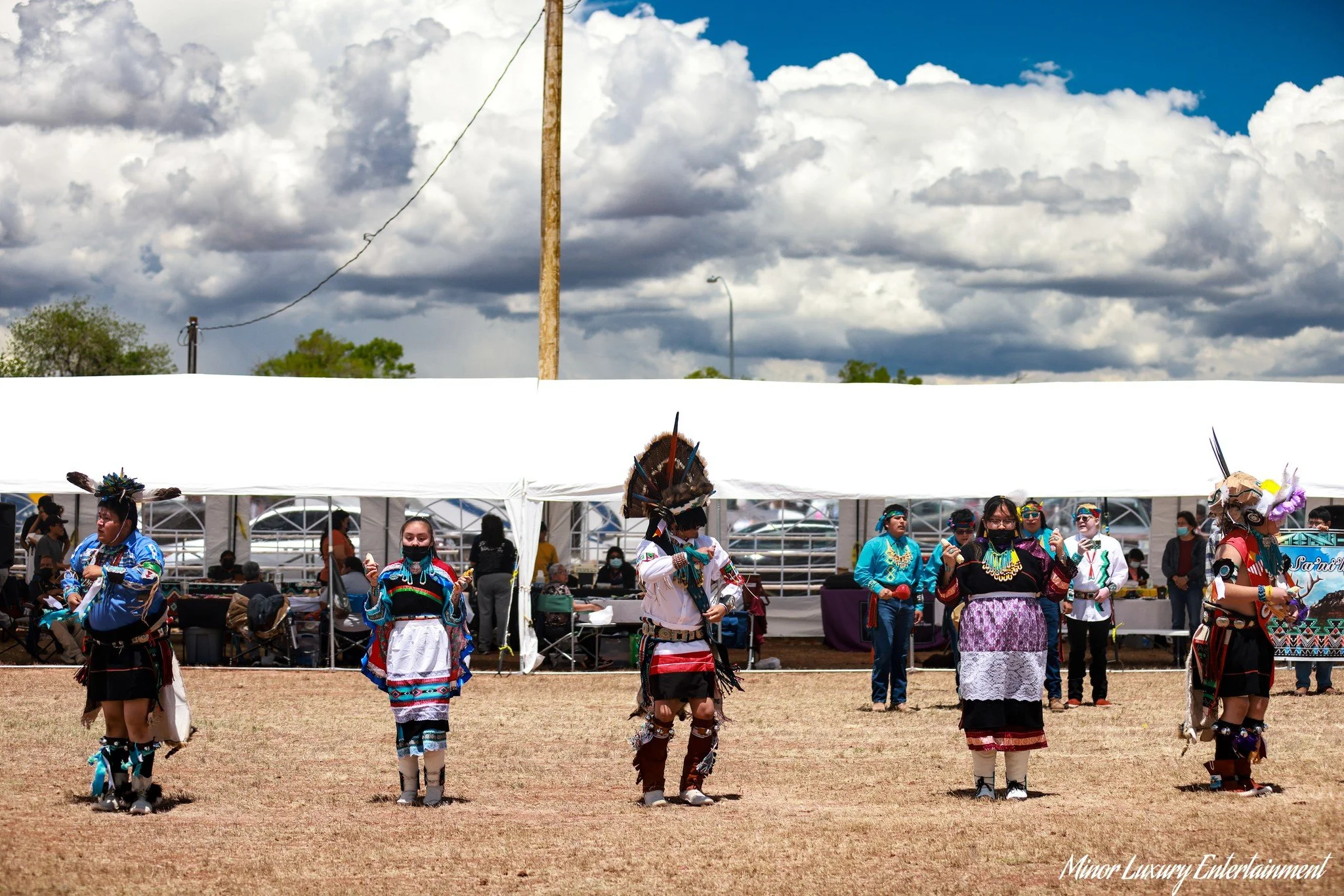 Group of Native American dancers in traditional attire performing outdoors under a partly cloudy sky, with onlookers and a white tent in the background.