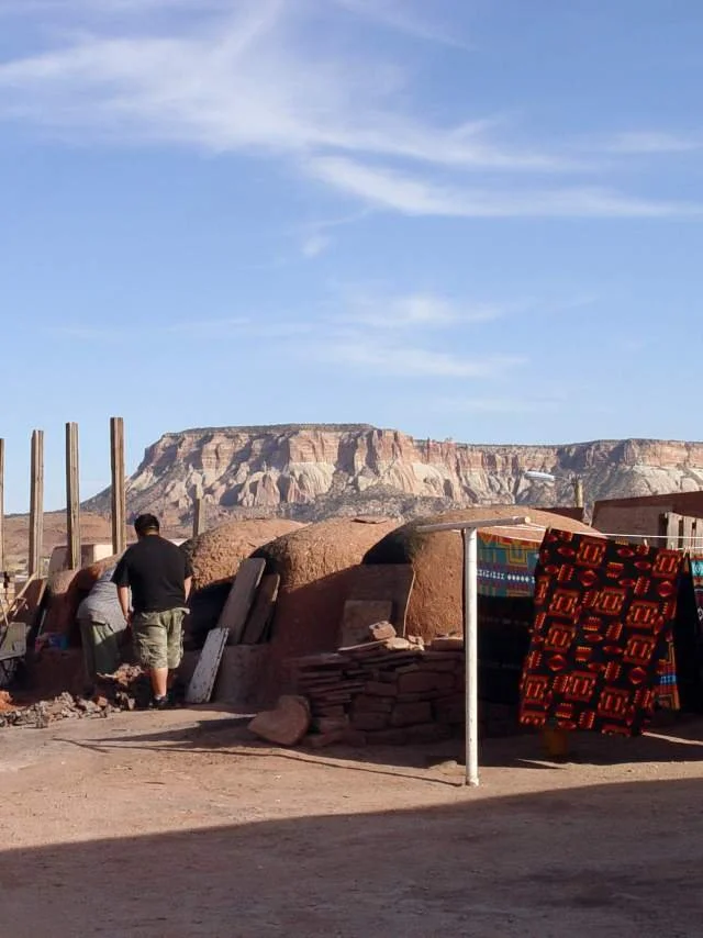 People walking in a desert landscape with rock formations. A clothesline with colorful woven textiles is visible in the foreground.