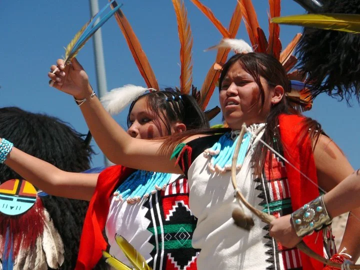 Two young women in traditional Zuni ceremonial attire, wearing feathered headdresses and necklaces, participating in a cultural dance outdoors.