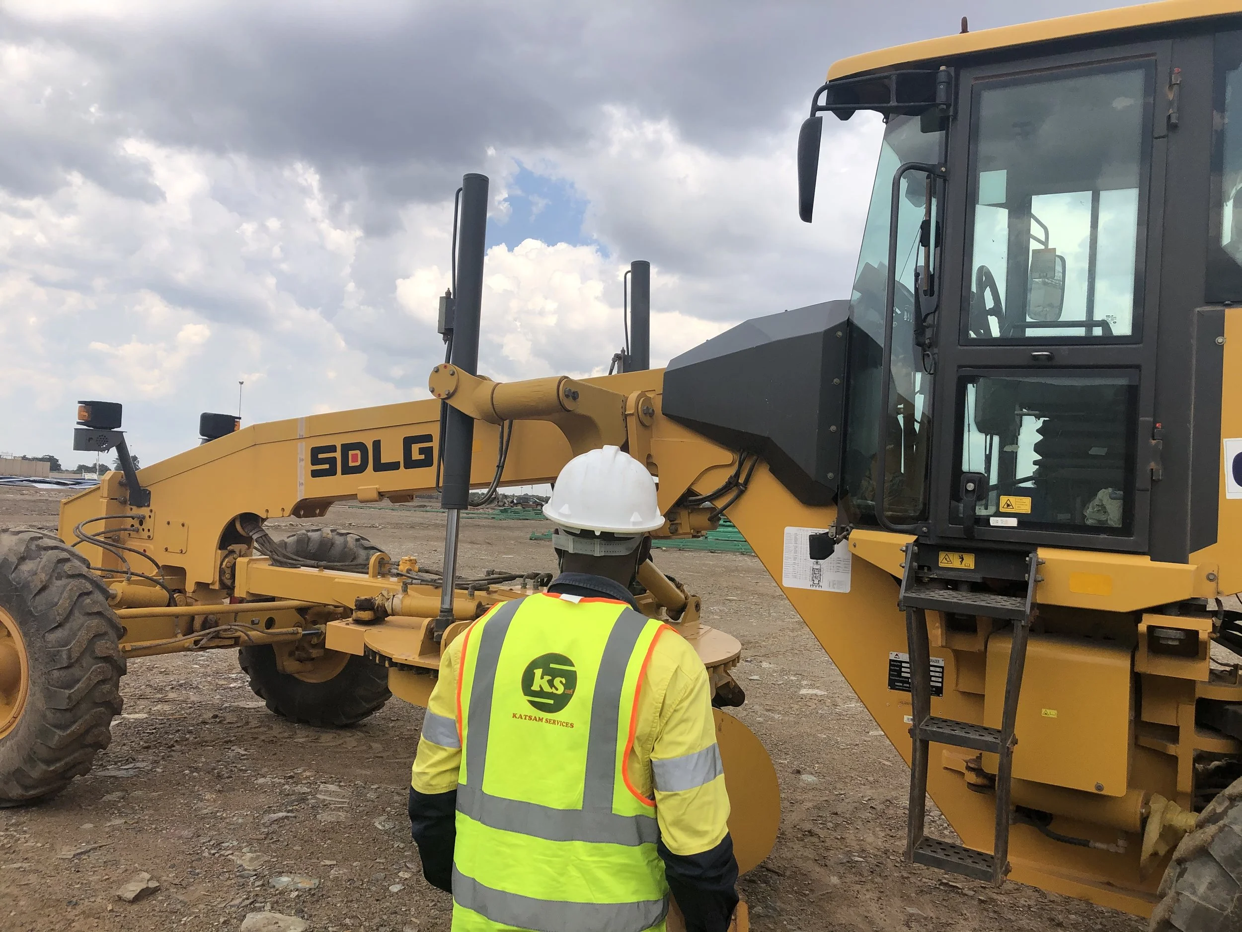 A construction worker in a high visibility yellow vest and white hard hat standing in front of a large yellow SDLG construction vehicle at a construction site with cloudy sky.