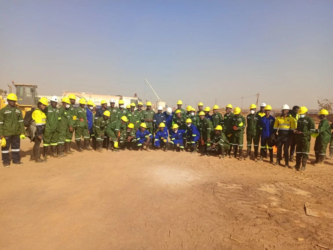 Group of construction workers and engineers wearing safety helmets and reflective clothing at a construction site with machinery and flat dirt ground.