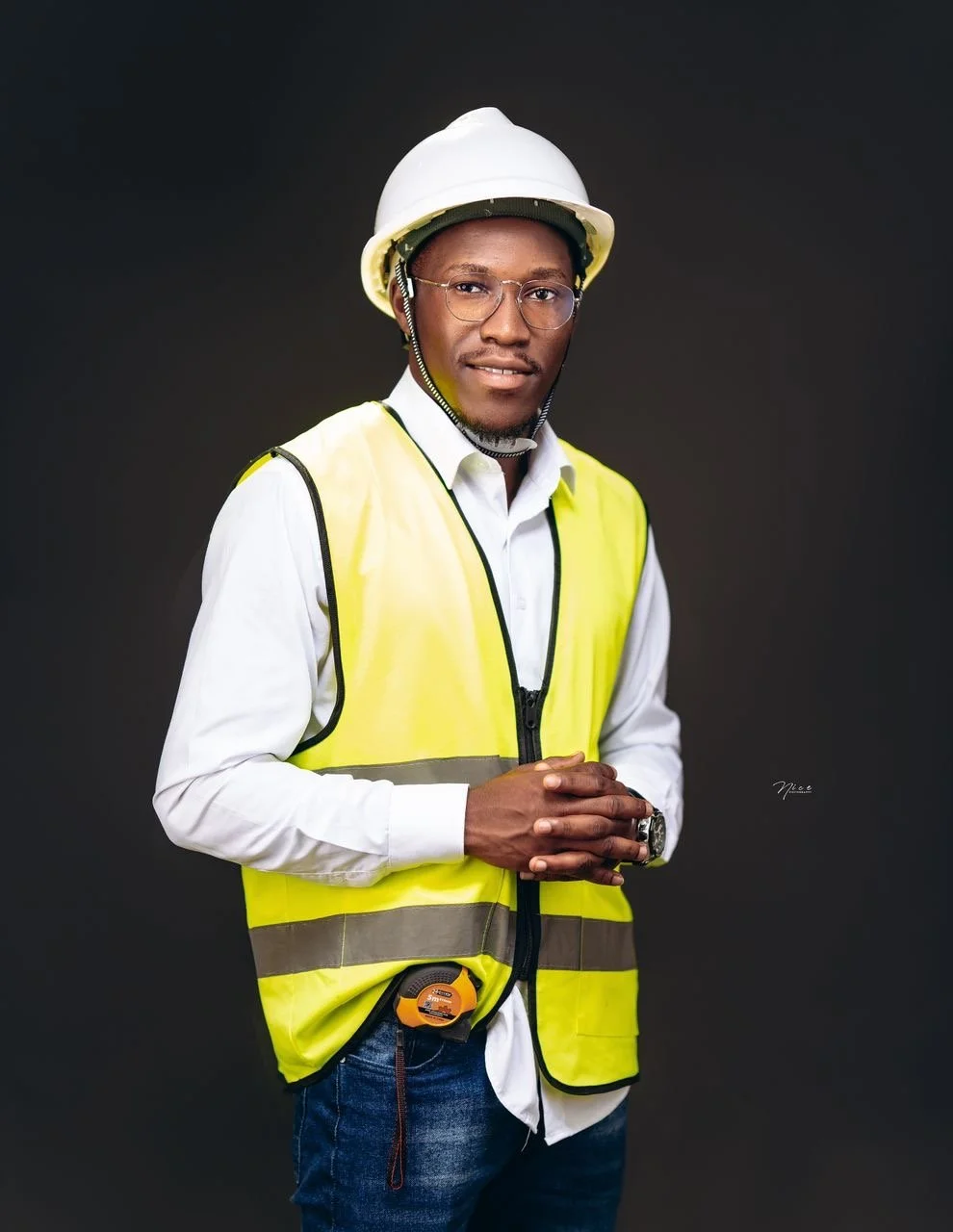 A young man wearing a white hard hat, clear glasses, a white shirt, and a yellow reflective safety vest, standing with hands clasped against a dark background.
