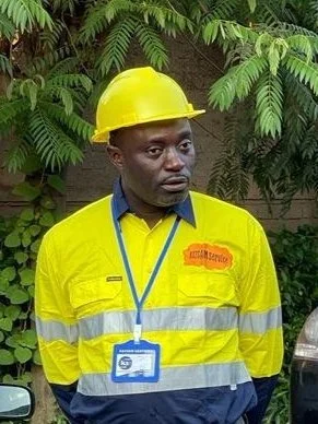 A man wearing a yellow safety helmet and a yellow high-visibility safety jacket with reflective stripes, standing outdoors with green foliage in the background.