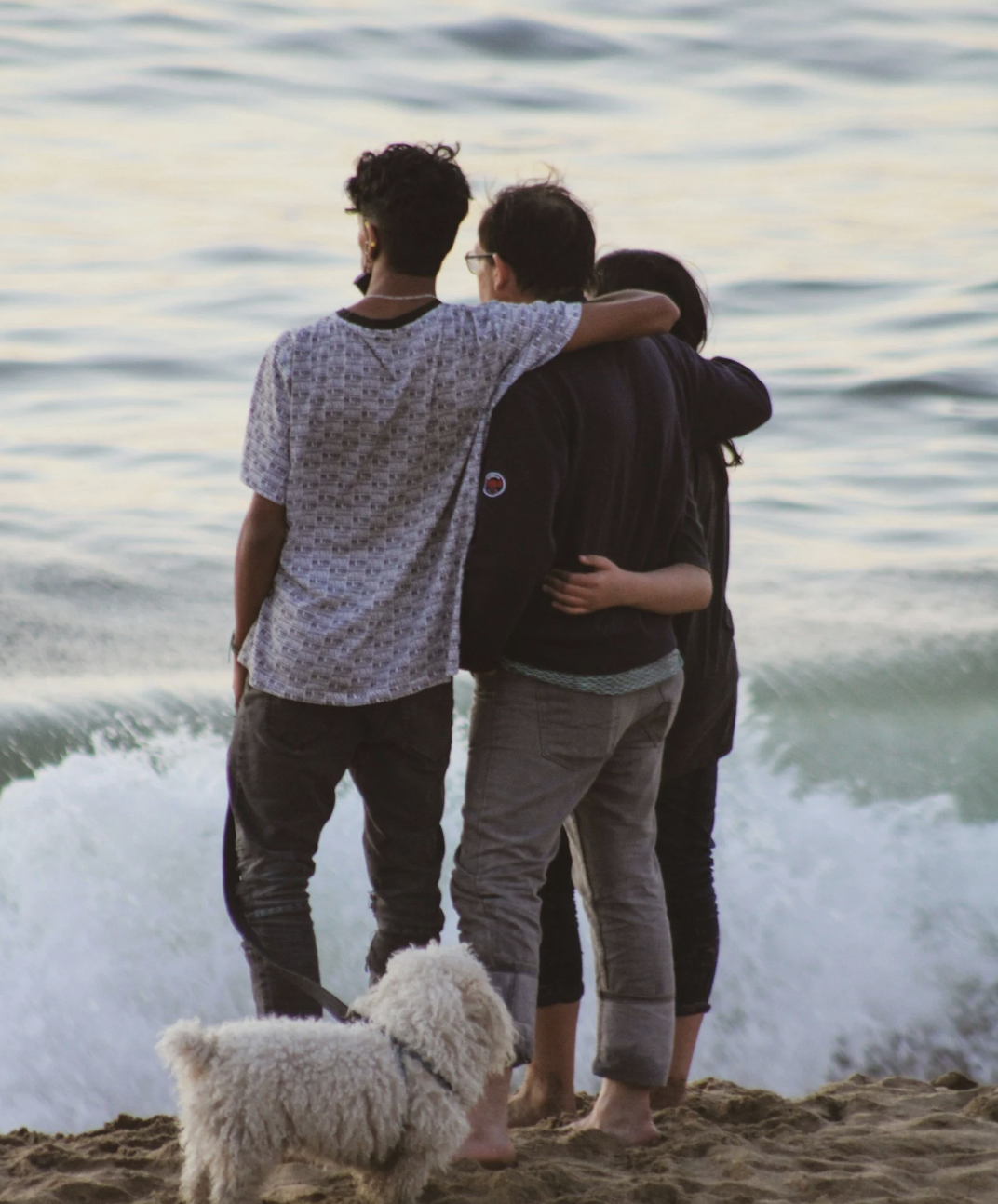 Three people standing on the beach near the ocean, with an adorable white fluffy dog. They are embracing and looking out at the water during sunset or early evening.