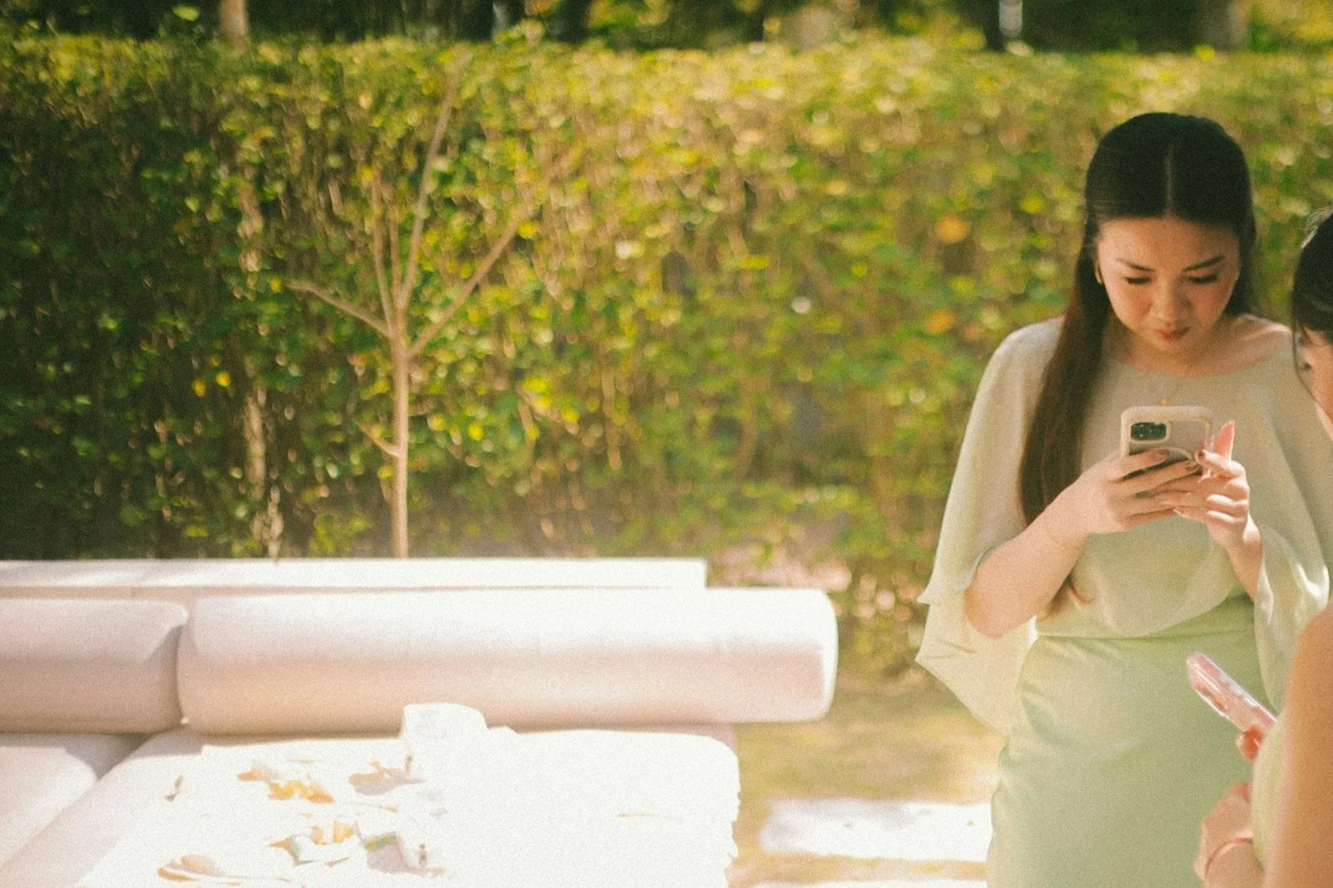 A woman in light-colored clothing looking at her phone outdoors at night. There is a white bench in front of her, and a background of green bushes and trees.