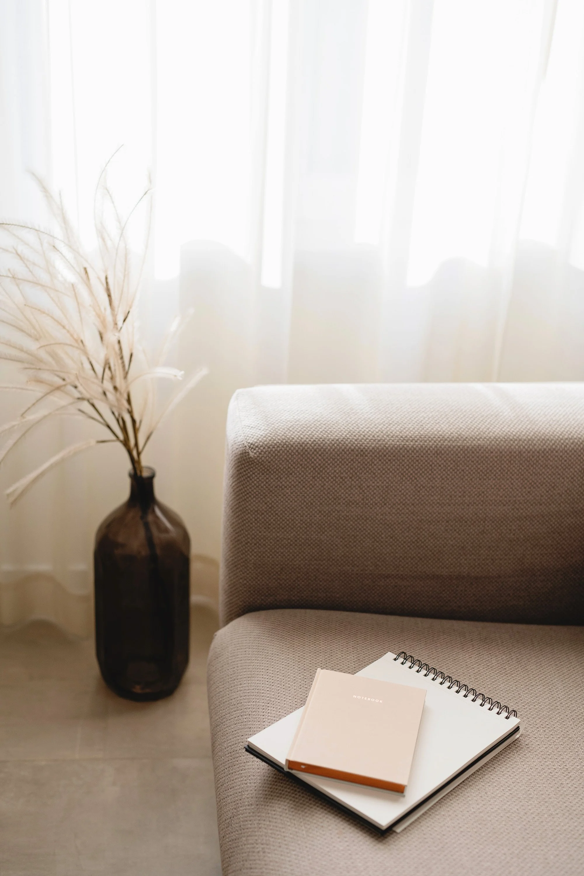 Interior of a room with a light-colored sofa, a dark vase with dried branches, a window with sheer white curtains, and a notebook with a pen on the sofa.