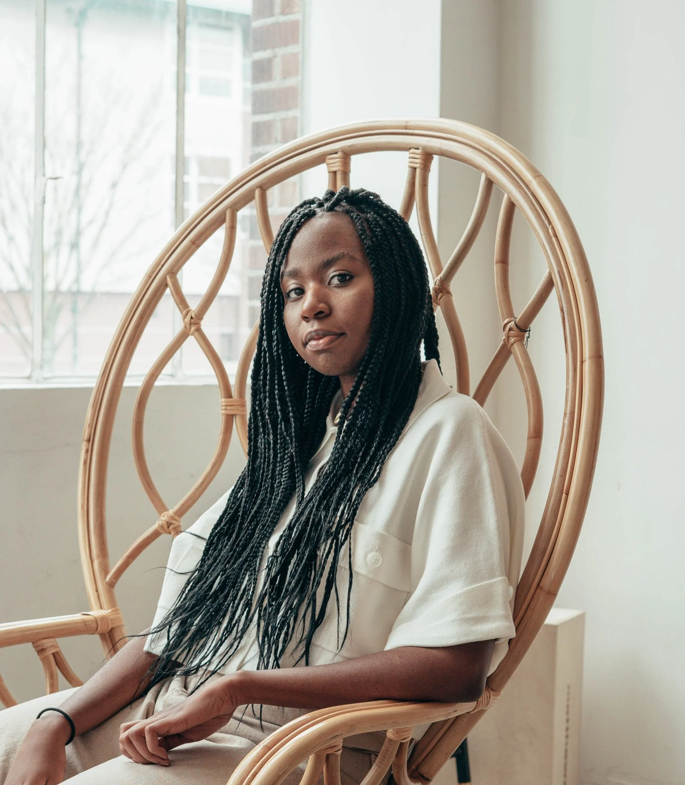 A woman with long braided hair sitting in a wicker chair near a window, with a neutral expression on her face.