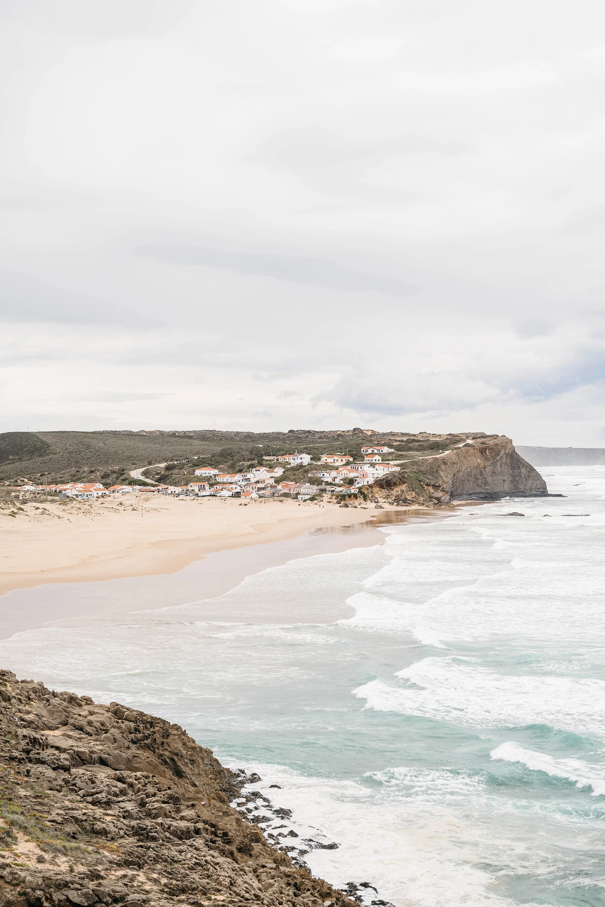 Overcast day at a coastal beach with rocky shoreline, white sandy beach, and waves. Residential houses on a hill in the background.