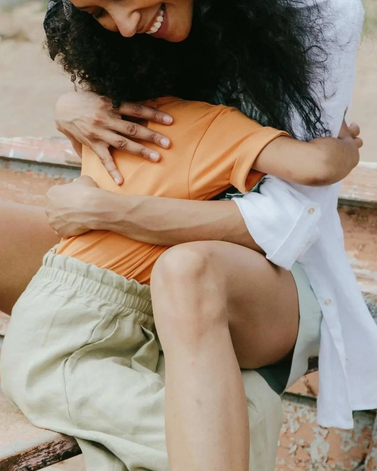 A person with long curly black hair is sitting on a step, hugging a child with light skin and short hair. The adult is smiling and leaning down, and the child is wearing an orange shirt and beige shorts. The background includes a brick wall and steps.