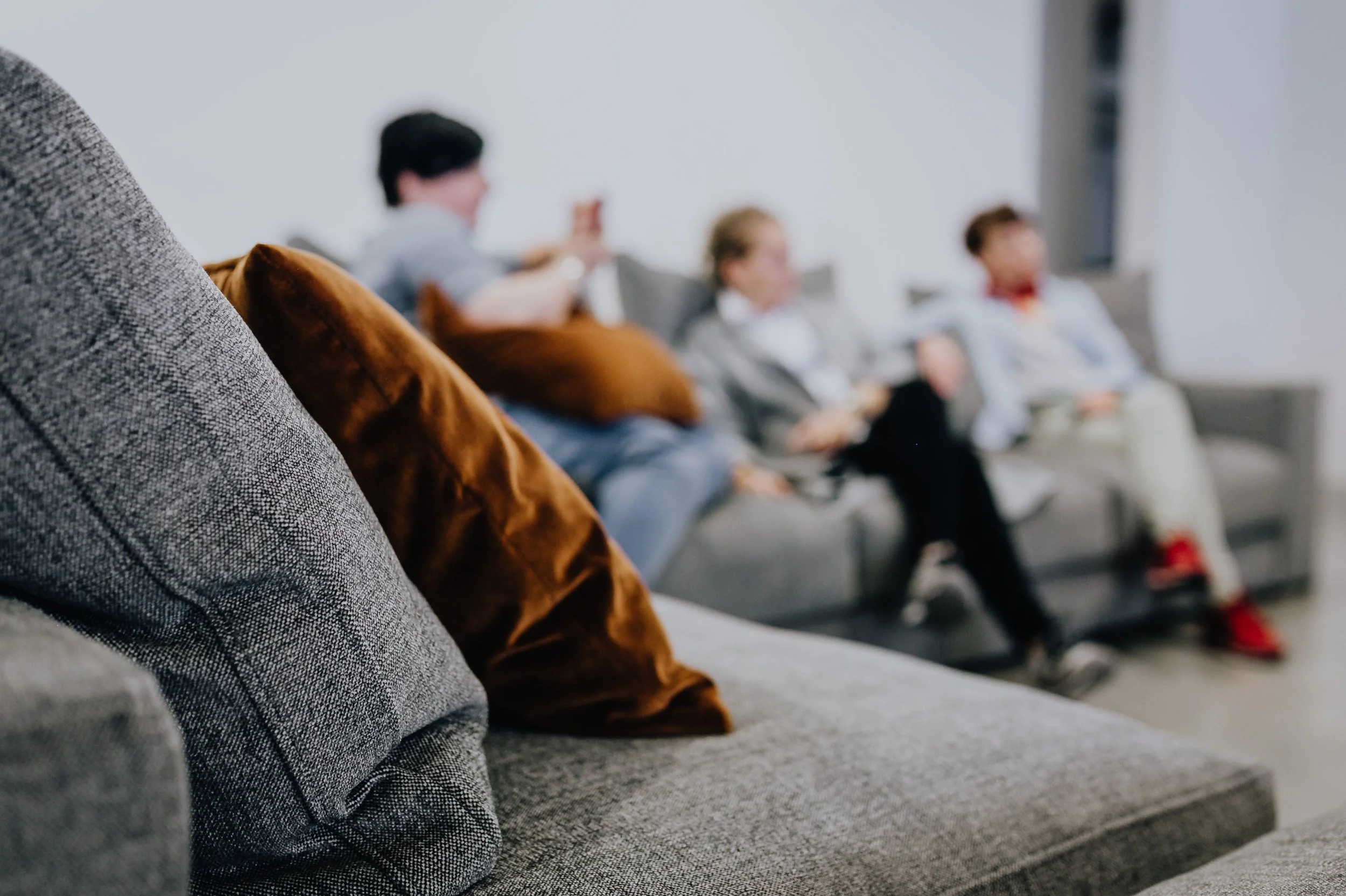 Close-up of a textured gray fabric on a piece of furniture in the foreground with three out-of-focus people sitting on a gray couch in the background, including a woman with blond hair and two men, one holding a phone.
