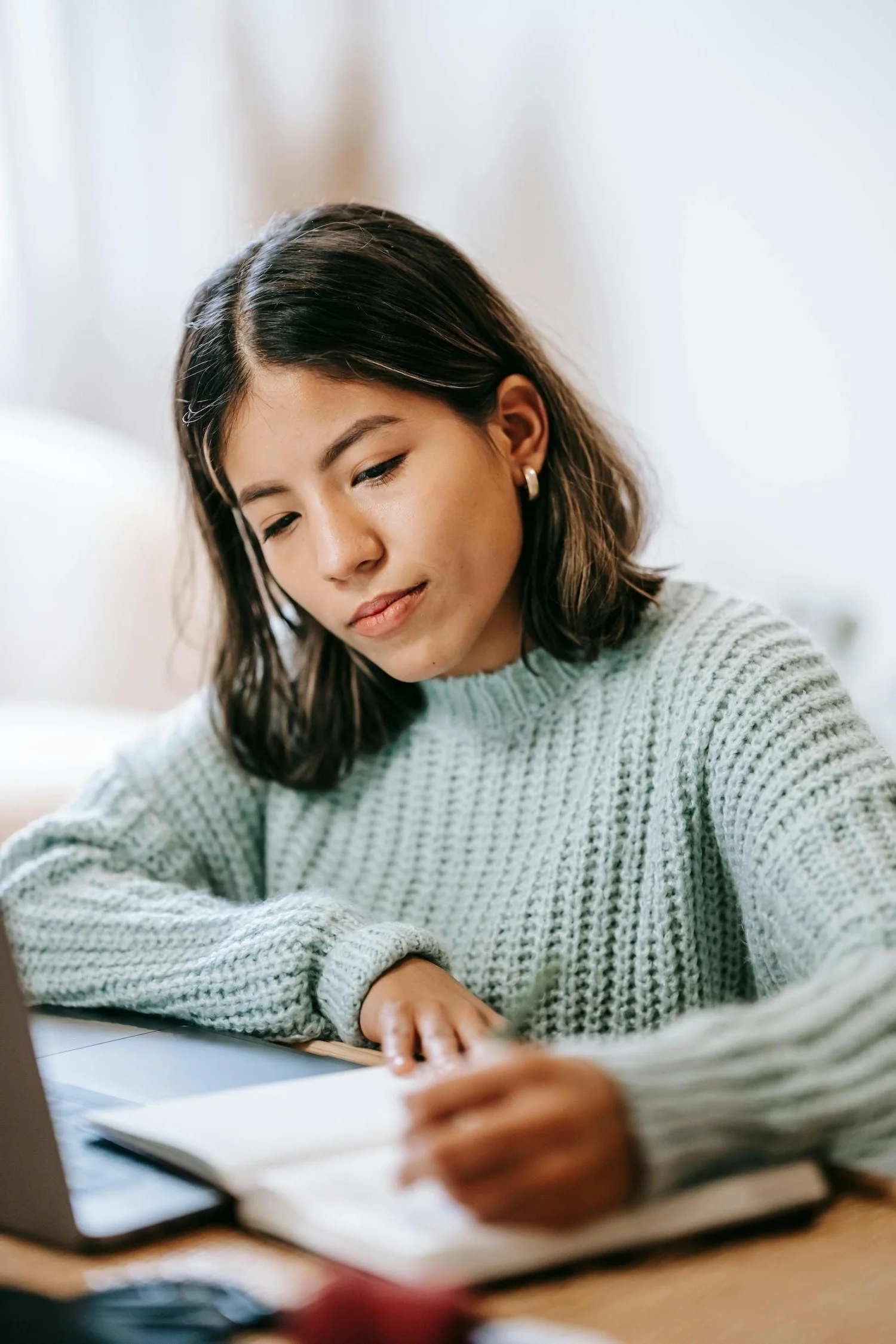 A woman with dark, shoulder-length hair wearing a light green, chunky knit sweater and pearl earrings, sitting at a desk, looking down at a notebook or paper with concentration.