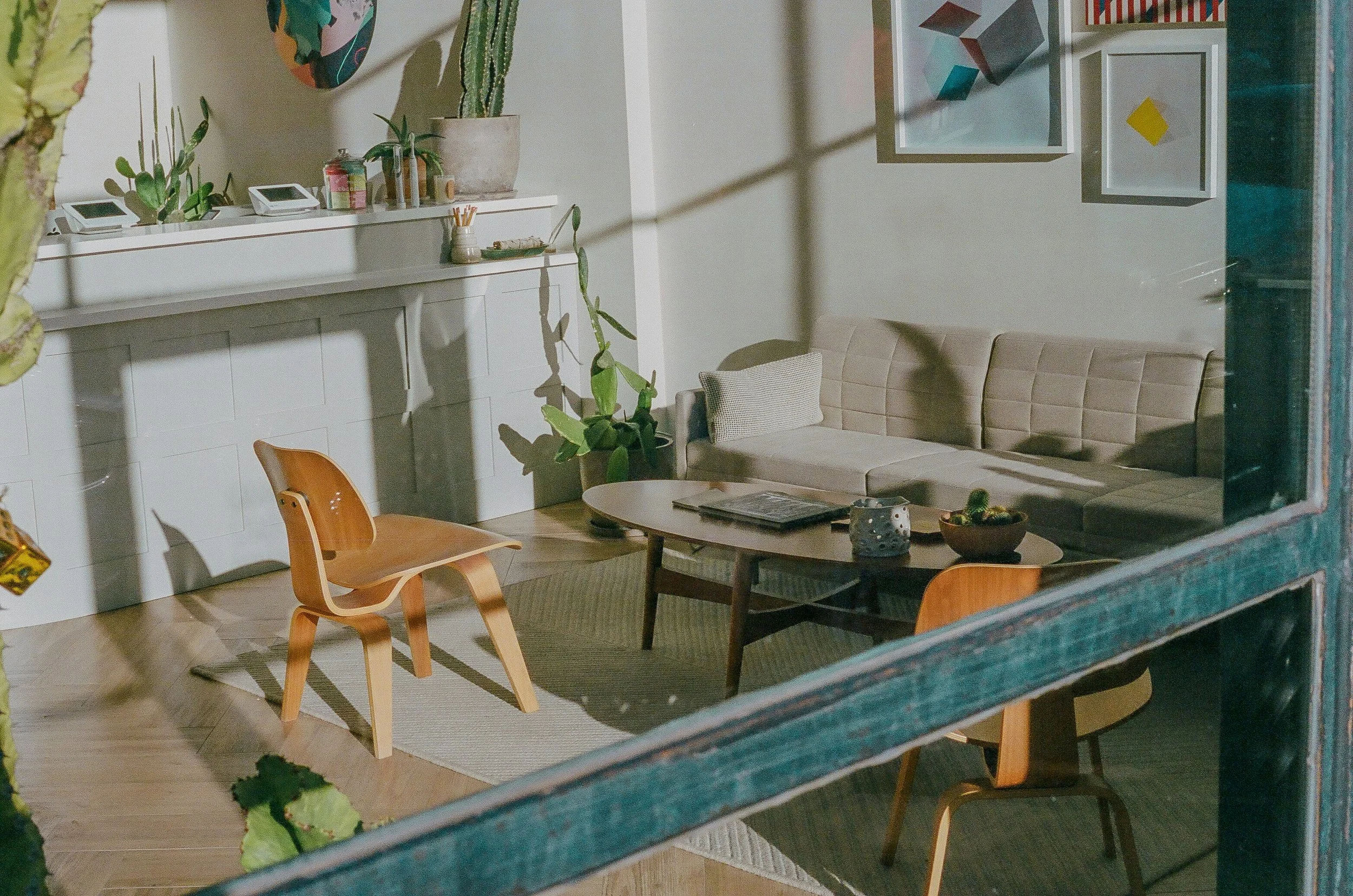 Living room seen through a window, with a beige sofa against the wall, a round wooden coffee table with magazines, a beige rug, a multicolored wall art, green plants including a cactus, and white cabinets with plants and objects on top.