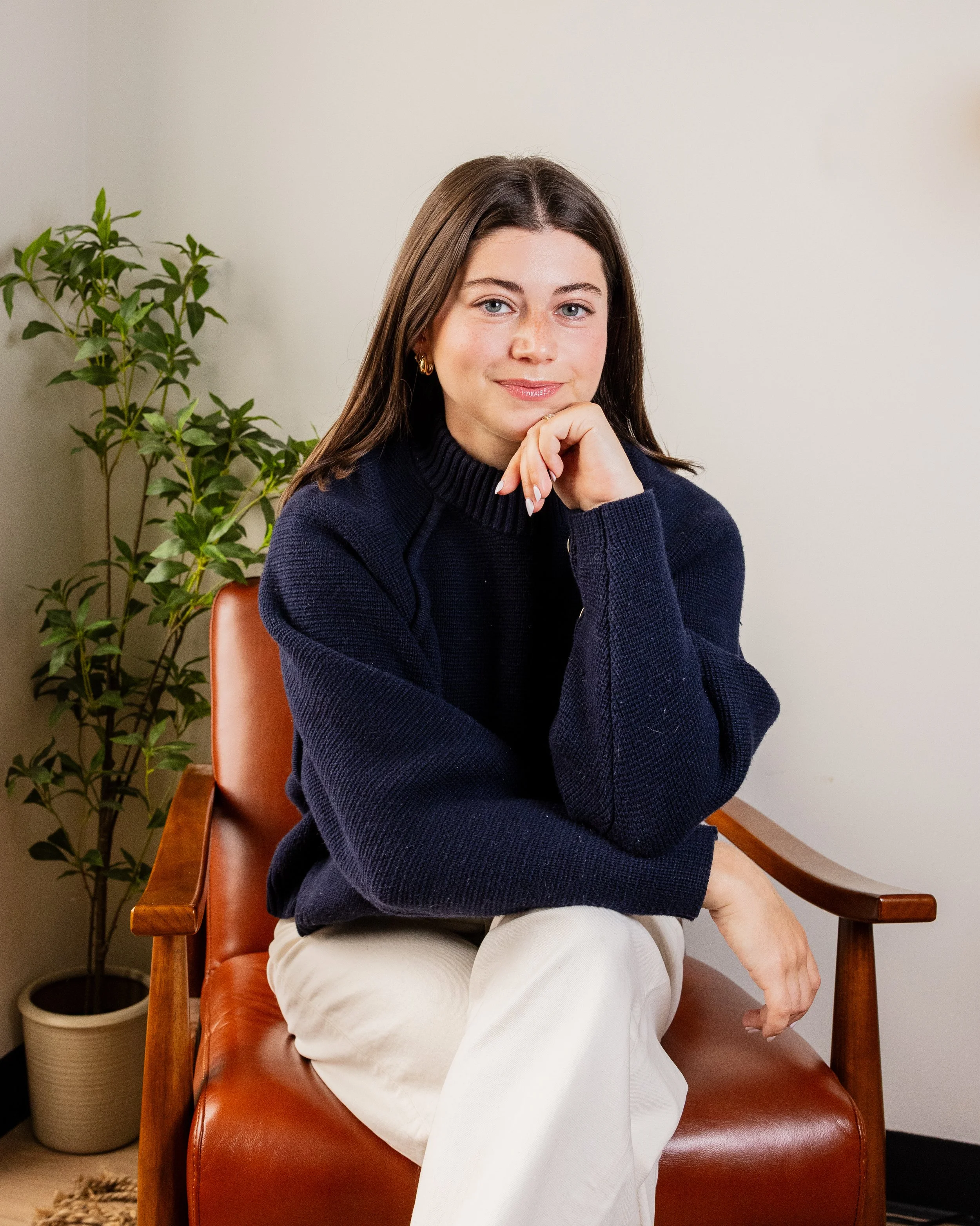 A woman with brown hair and blue eyes wearing a dark sweater sits on a wooden chair with a leather seat, resting her chin on her hand, with a plant and white wall in the background.