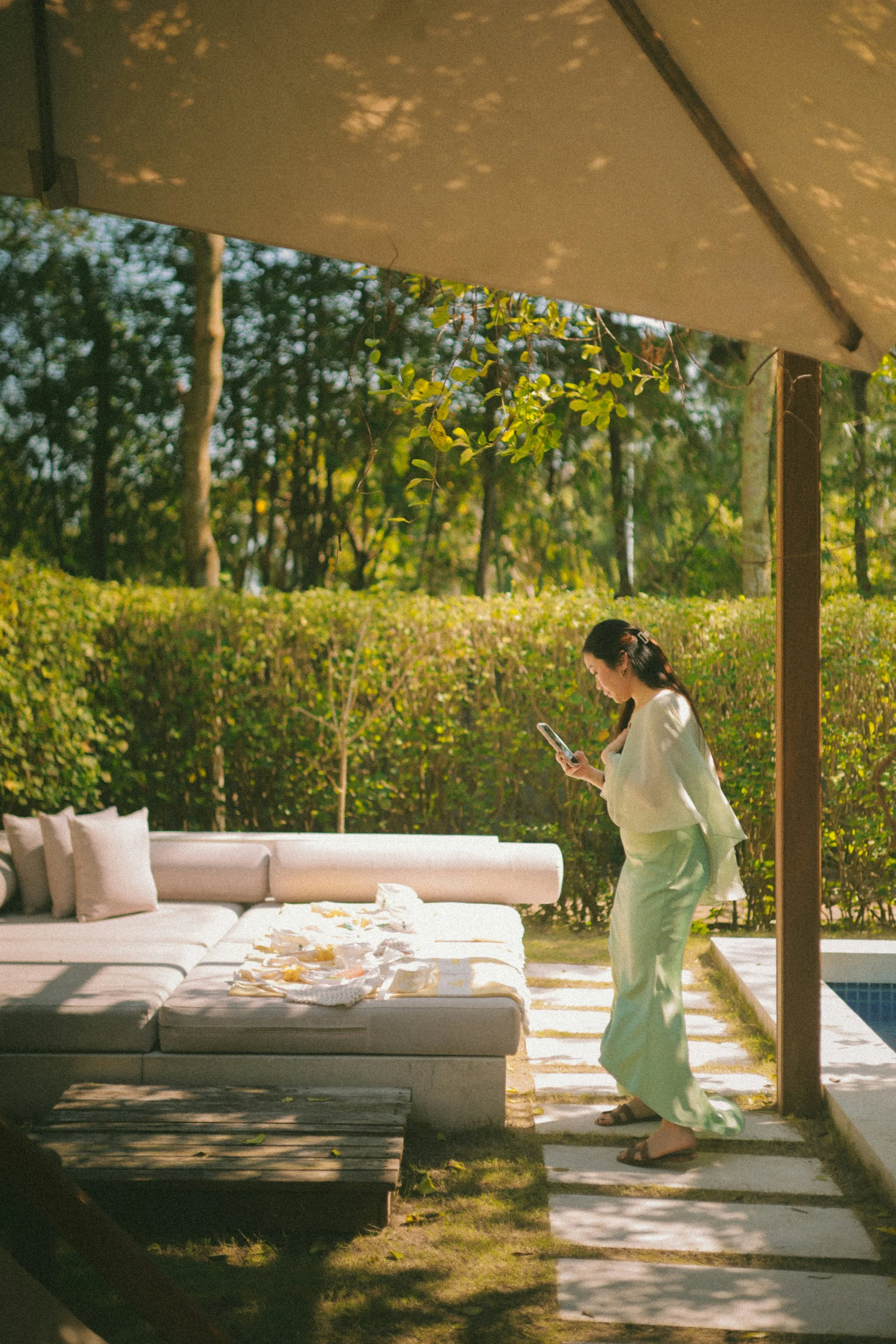 A woman in white pajamas is standing outdoors under a beige umbrella, looking at her phone. There is a white outdoor sofa with pillows and a table with food on it. The background features trees and greenery.