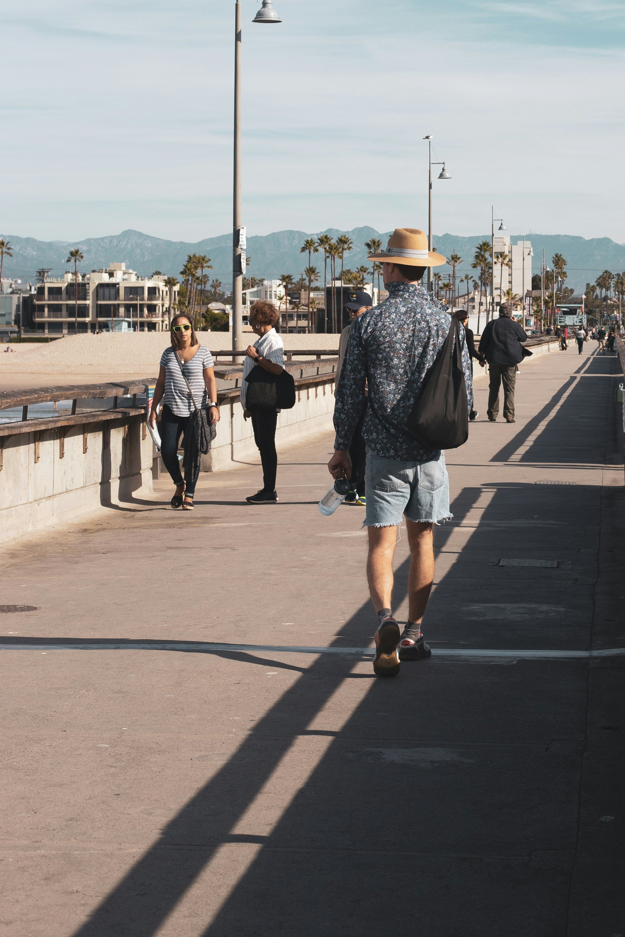 People walking along a concrete pathway near the beach with palm trees, mountains in the background, and a partly cloudy sky.