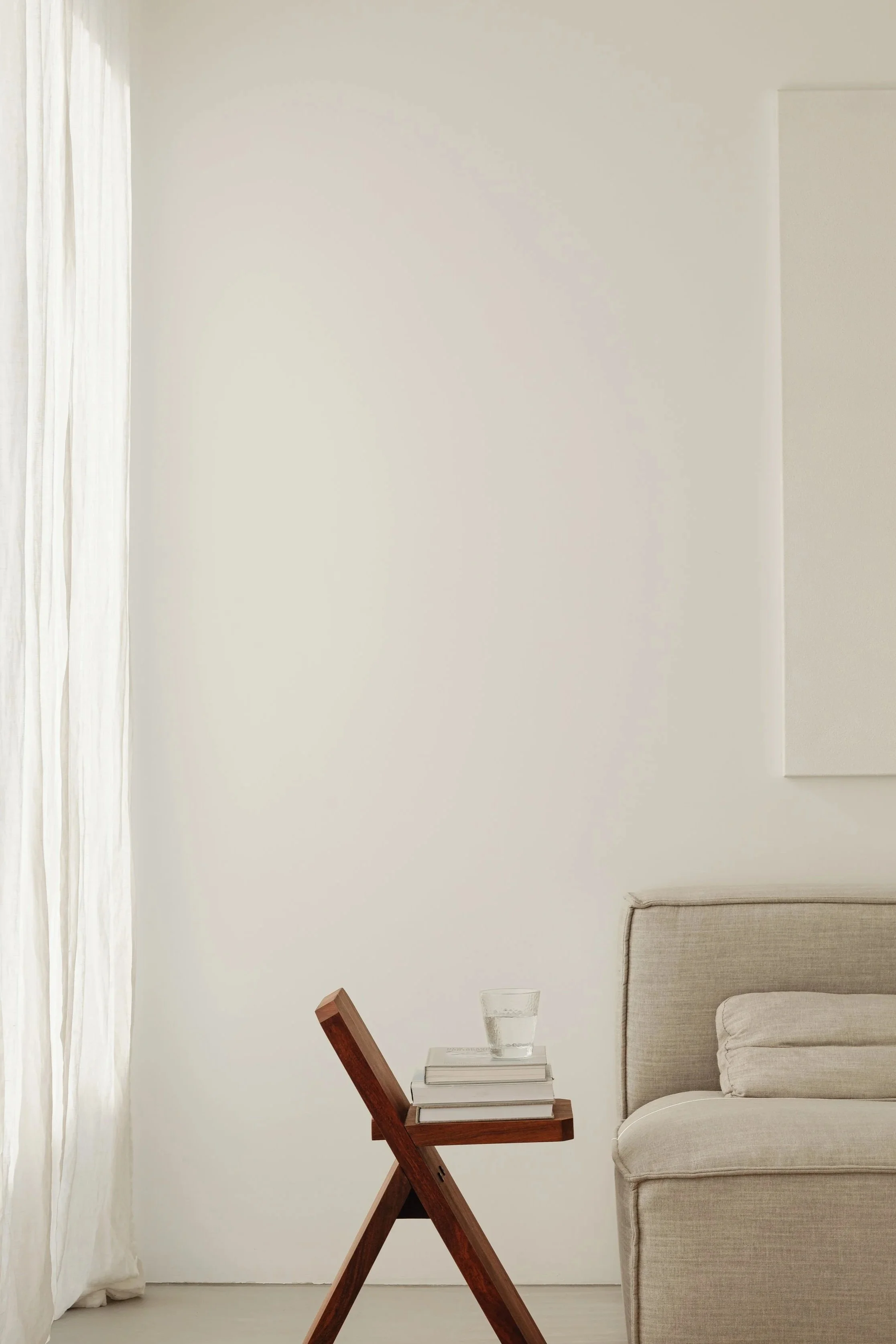 A minimalist living room corner with a beige sofa, a small wooden side table with books and a glass of water, cream curtains, plain cream wall, and a small decorative object on the floor.