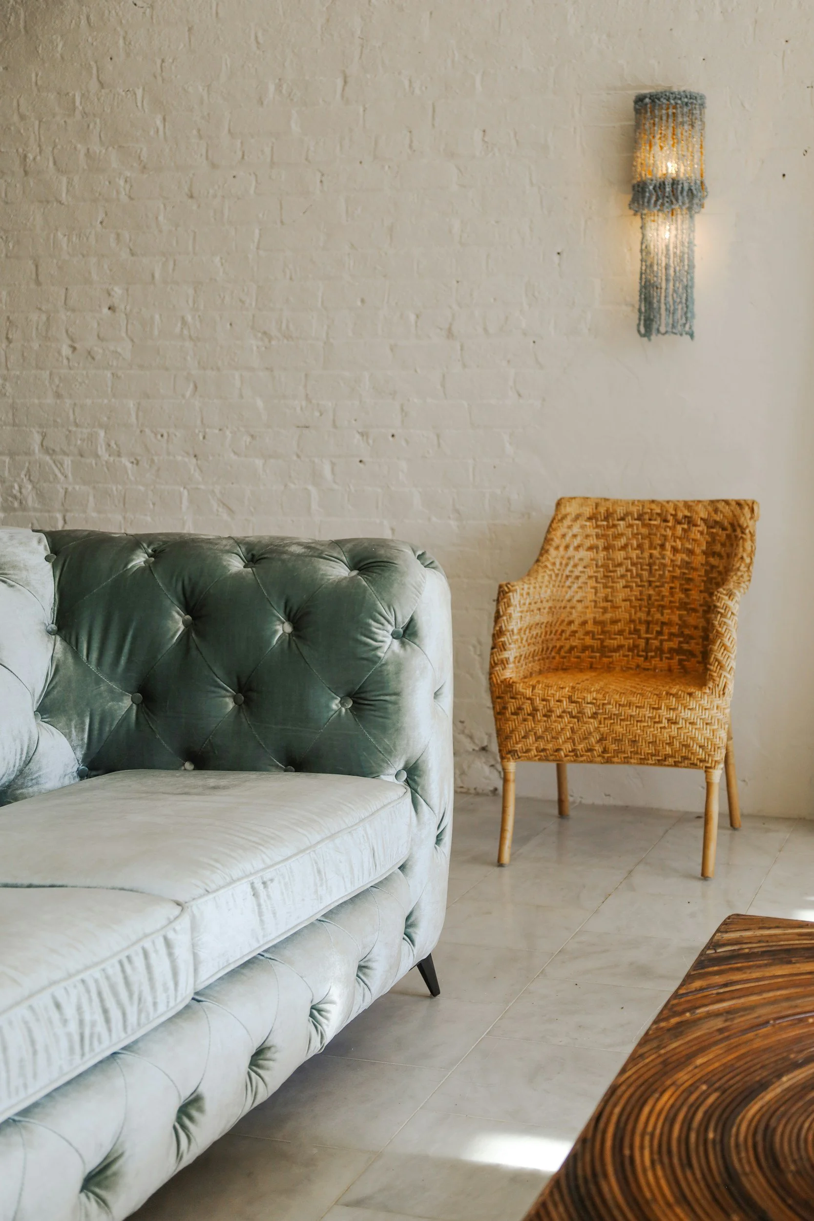 A light green tufted velvet sofa, a yellow woven chair, and a wooden table in front, against a white brick wall with a wall light fixture.