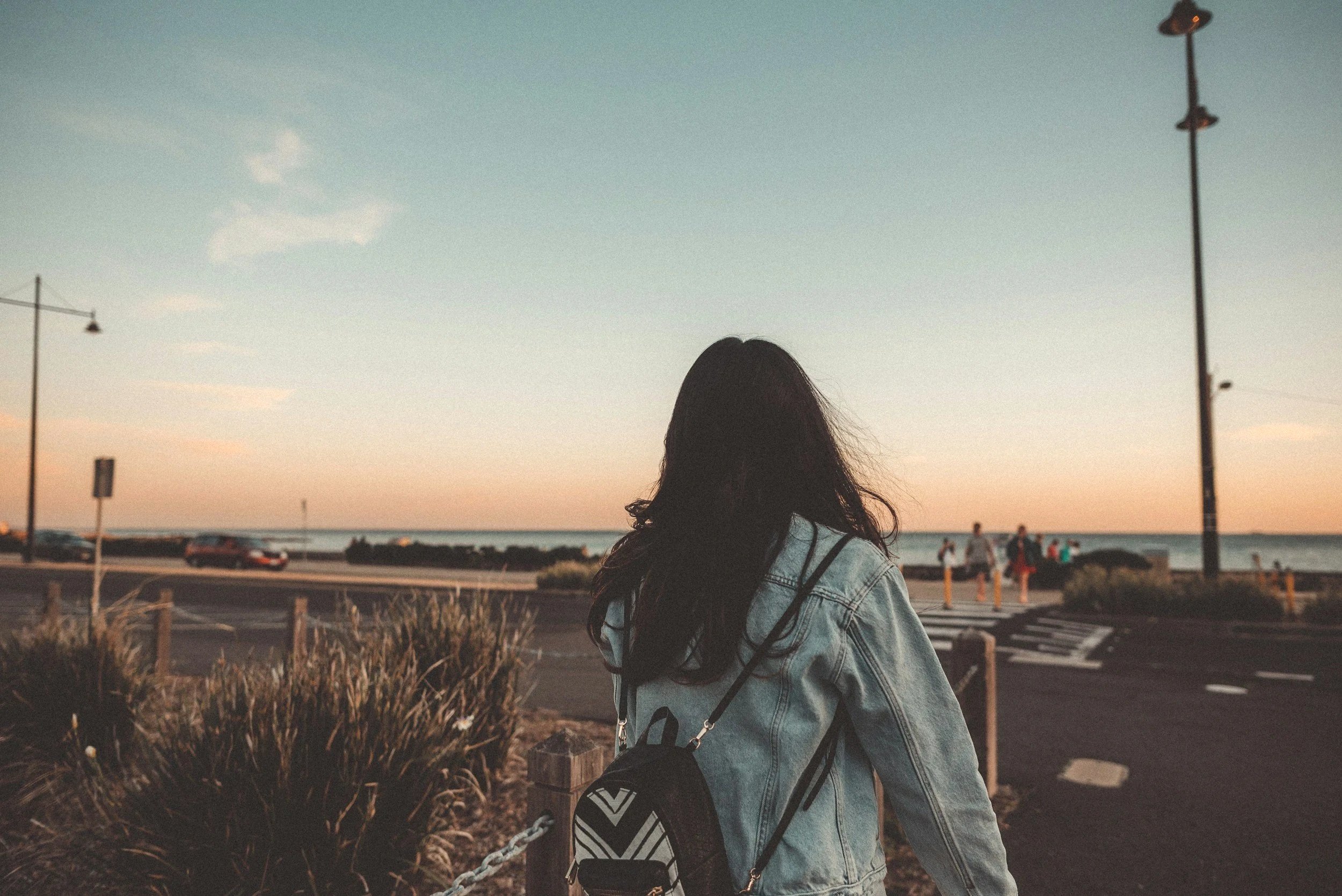Person with long hair wearing a denim jacket and carrying a patterned bag, viewed from behind, looking towards the sunset at a beach parking lot with streetlights and a few people in the background.