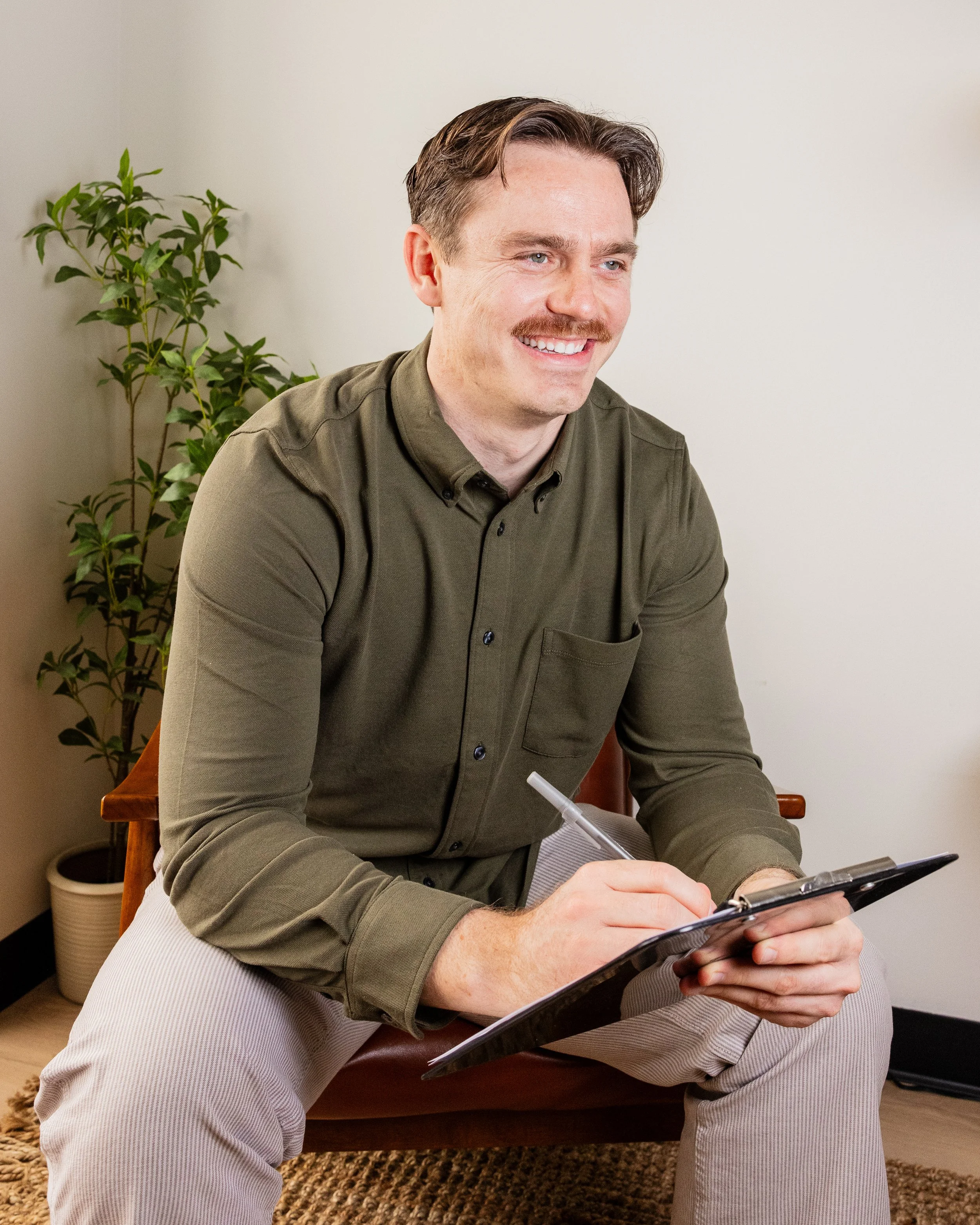 A man sitting on a chair, smiling, holding a clipboard and pen, wearing a green shirt and beige pants, with a plant in a white pot behind him in a room with beige carpet and white walls.