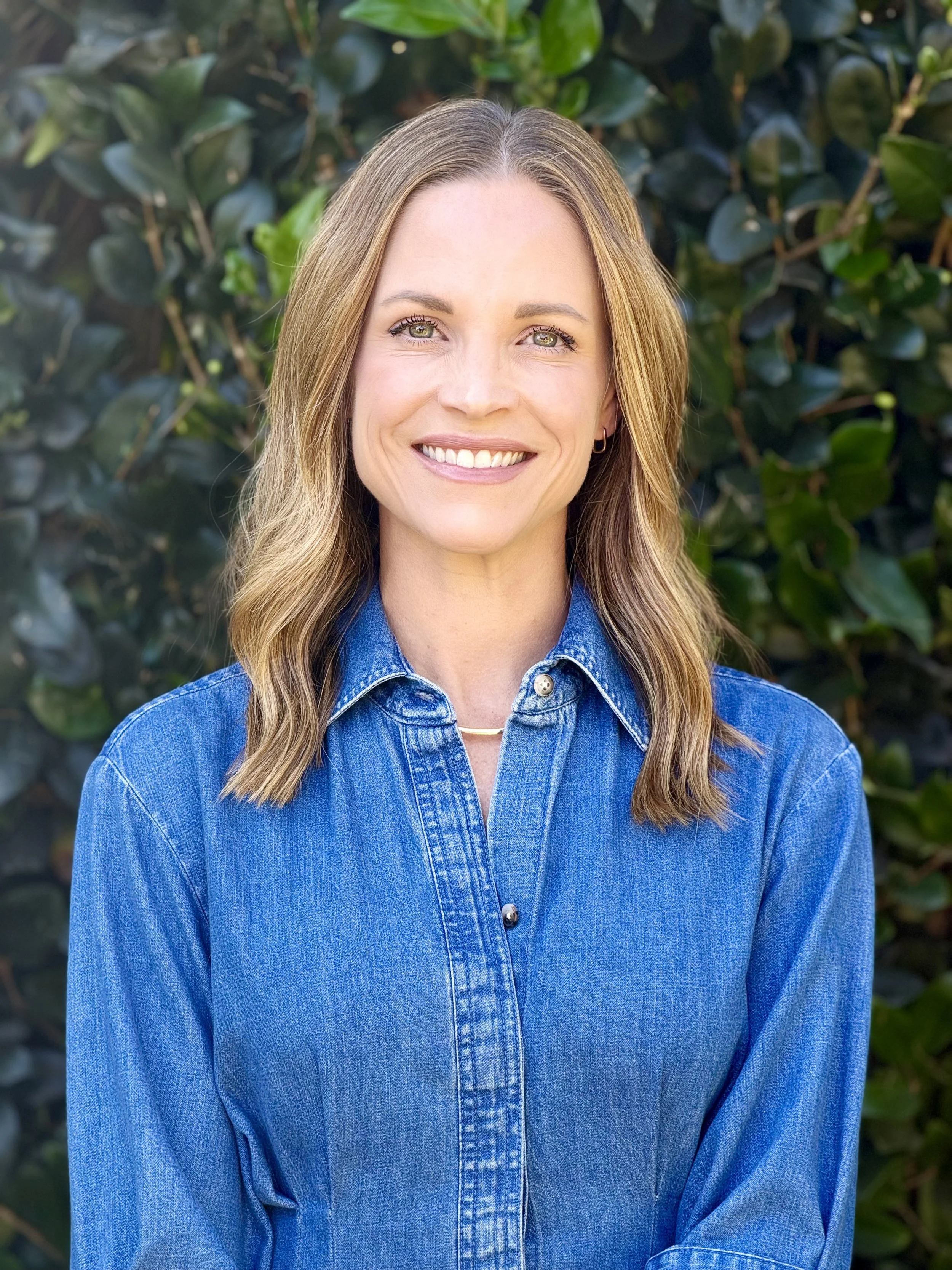 A woman with light brown hair, light skin, and green eyes smiling, wearing a blue denim shirt, standing outdoors in front of a leafy green hedge.
