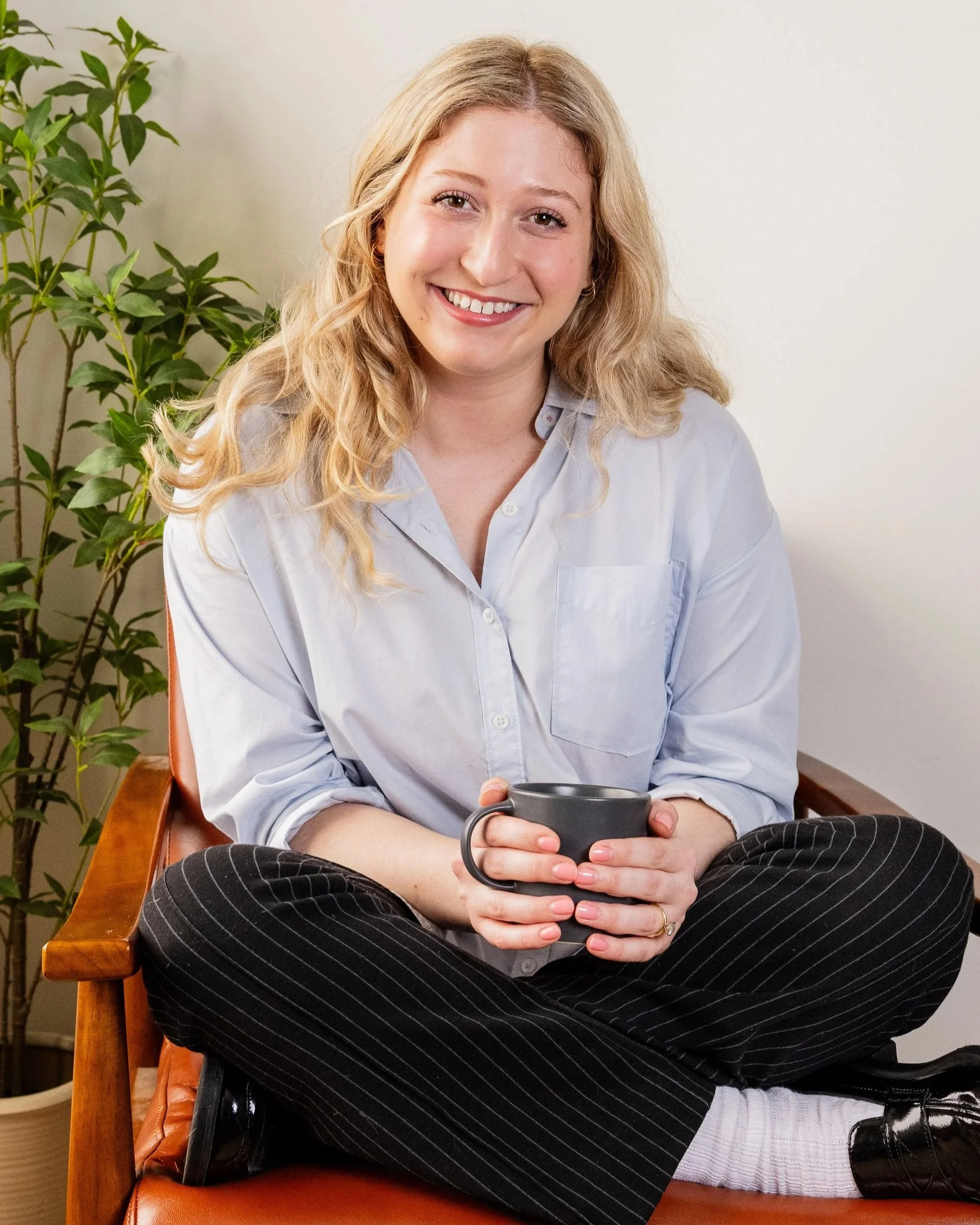 A woman with long blonde hair, wearing a light blue button-up shirt and black pinstripe pants, sitting on a wooden chair with a brown leather cushion, holding a gray coffee mug, smiling at the camera, with a green plant in the background.