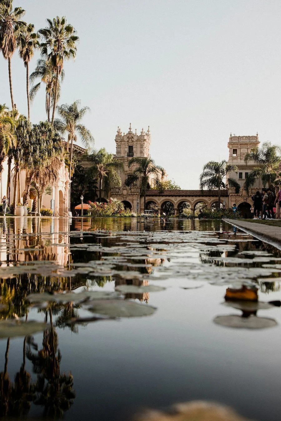View of a historic Spanish-style building with arches, towers, and ornate detailing, surrounded by palm trees, with a reflecting pool in the foreground and people walking nearby.